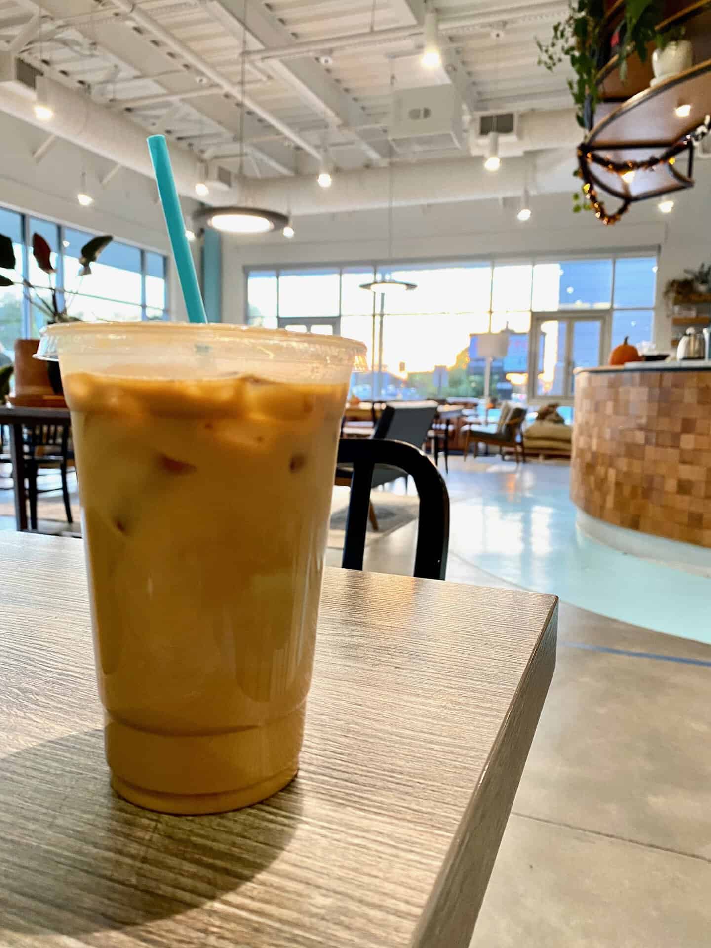 Iced coffee in a clear plastic cup with a blue straw on a wooden table in a modern cafe setting.