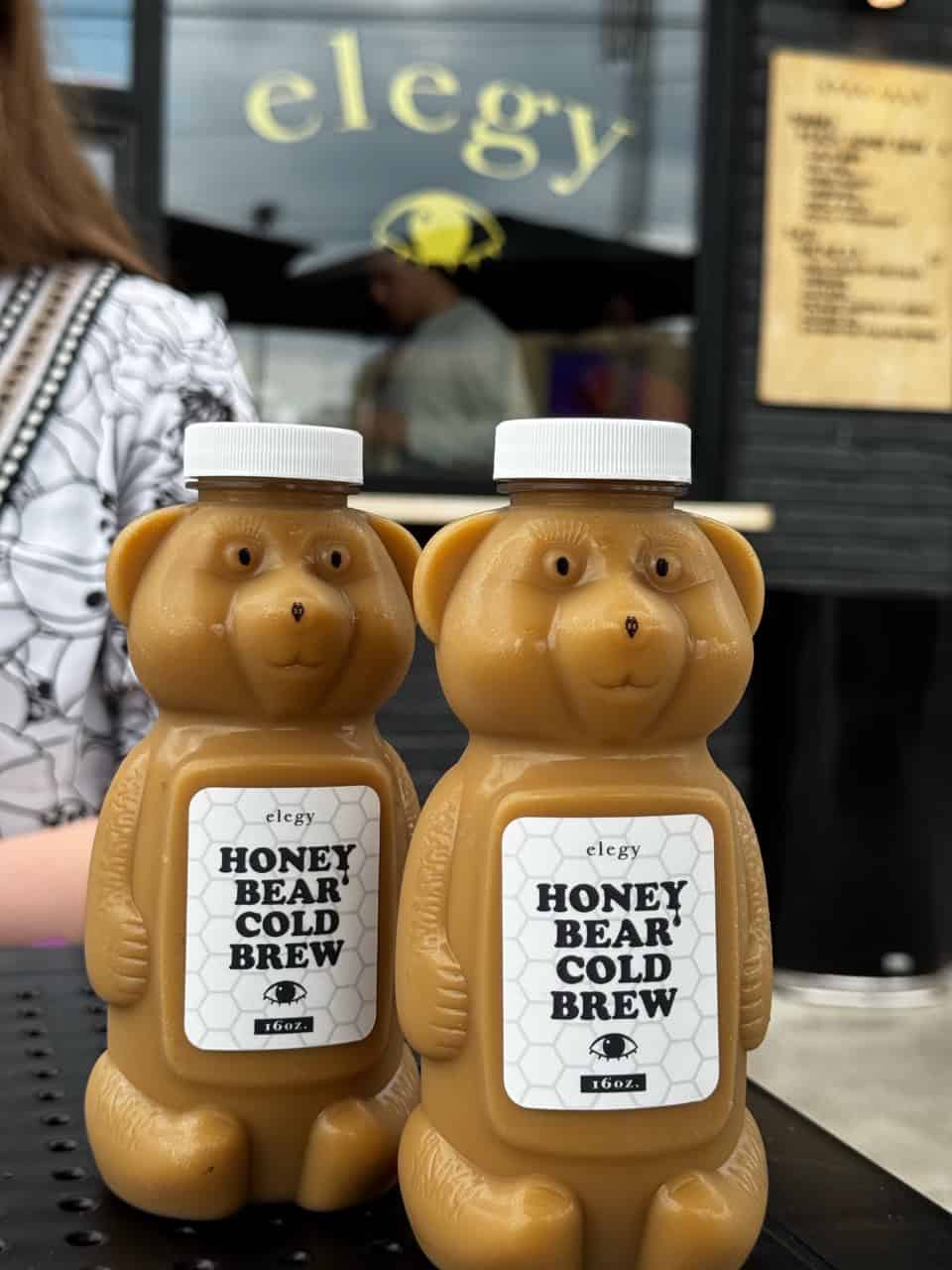 Two plastic bear-shaped bottles labeled "Honey Bear Cold Brew" rest on a table. In the blurred background, a sign reads "elegy," subtly capturing the Nashville vibe.