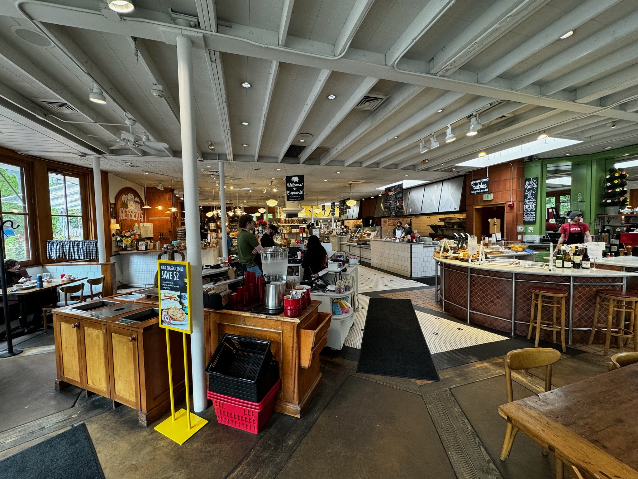 Spacious café interior with counters, display cases, and various seating areas. Customers and staff are visible, with food and drink stations throughout the space.