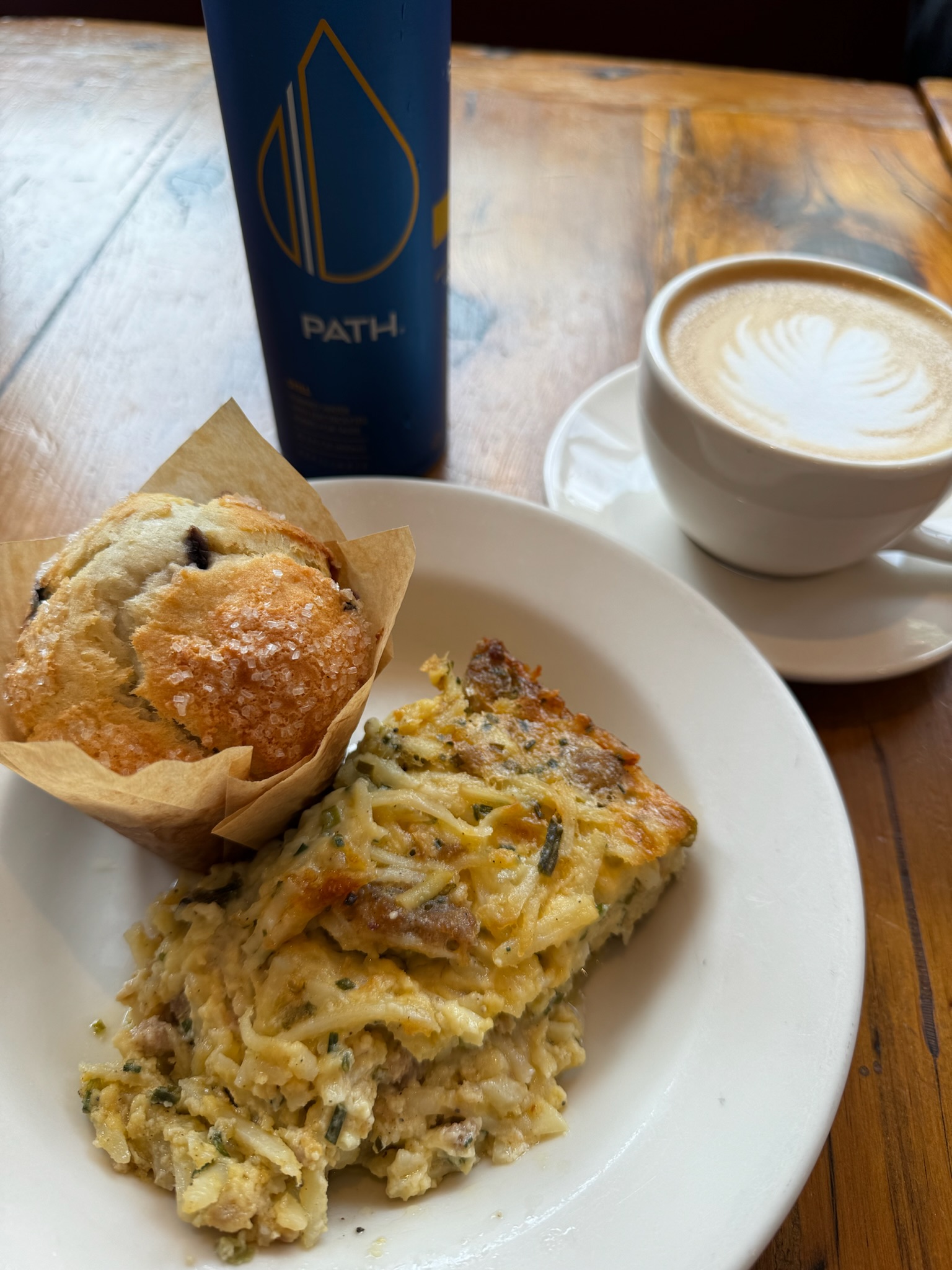 Plate with a slice of savory casserole and a blueberry muffin, next to a cup of latte with foam art and a blue PATH water bottle on a wooden table.