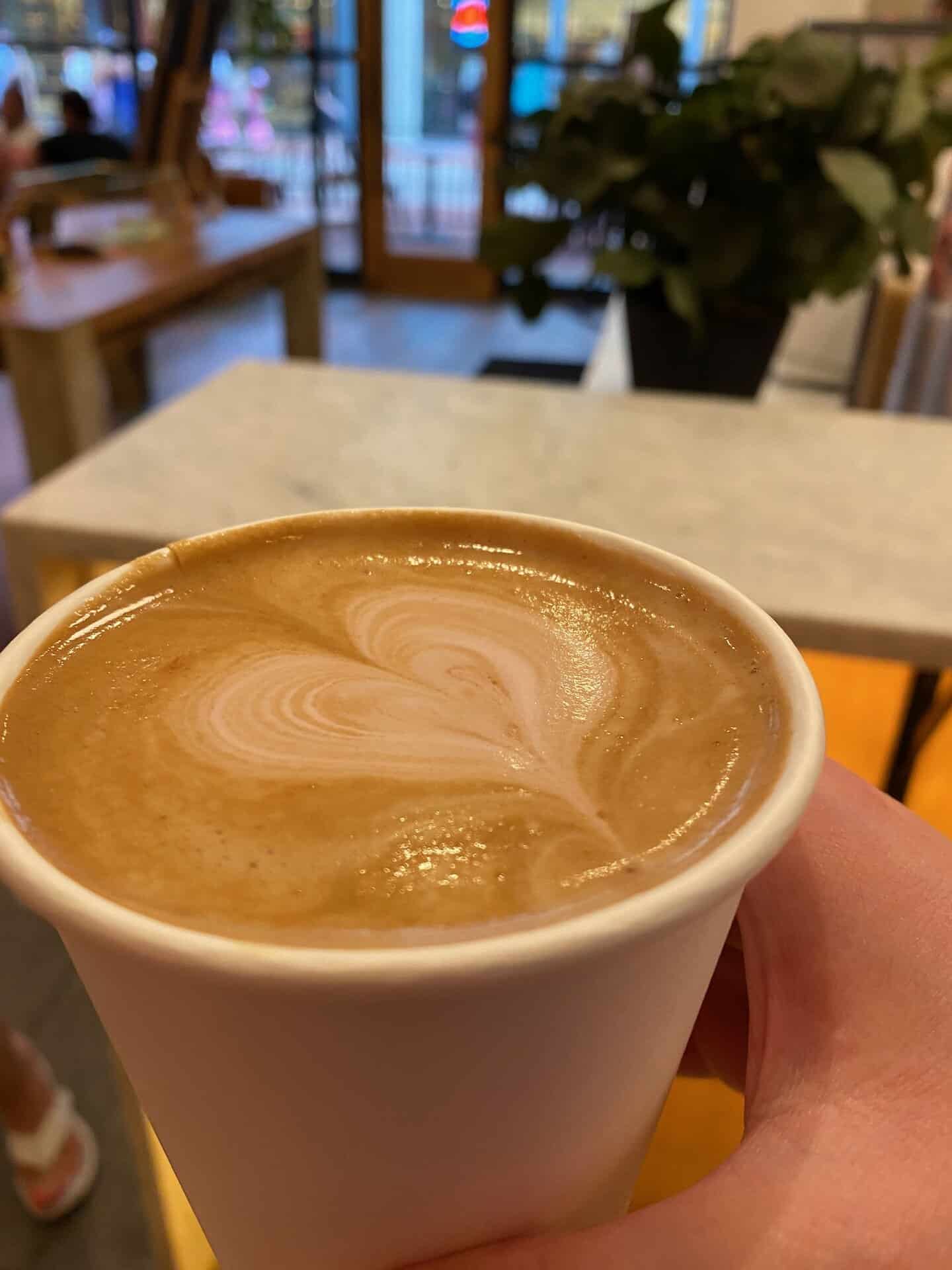 A hand holding a paper cup of coffee with heart-shaped latte art, inside a café with blurred background details.