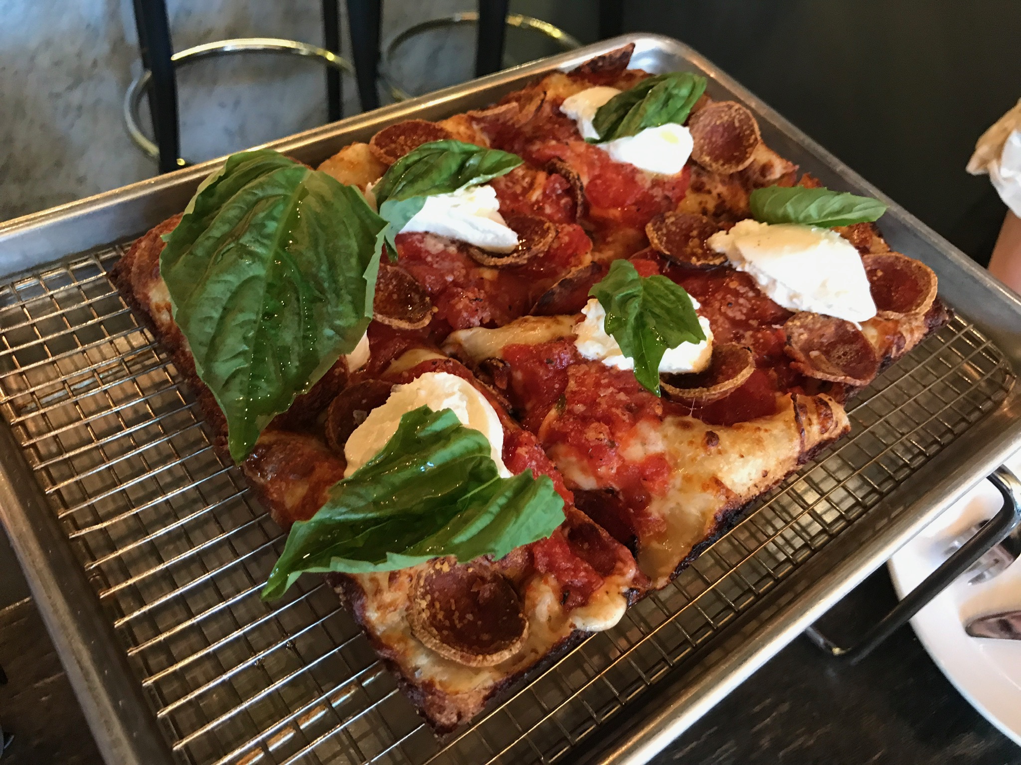 Square slices of pepperoni pizza topped with dollops of ricotta cheese and fresh basil leaves are served on a wire rack over a metal tray.