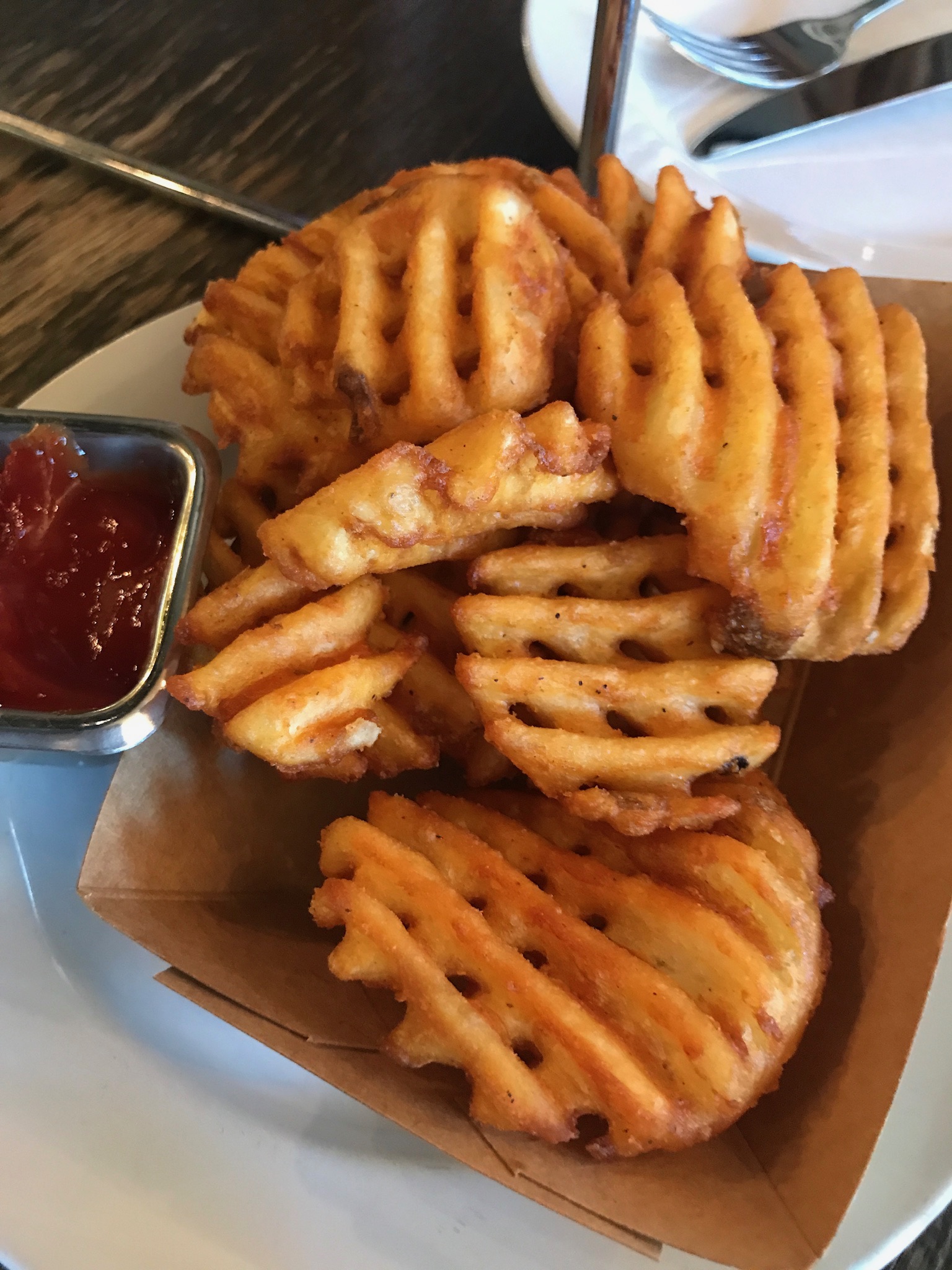 Waffle fries served in a brown paper tray with a small metal container of ketchup on the side.