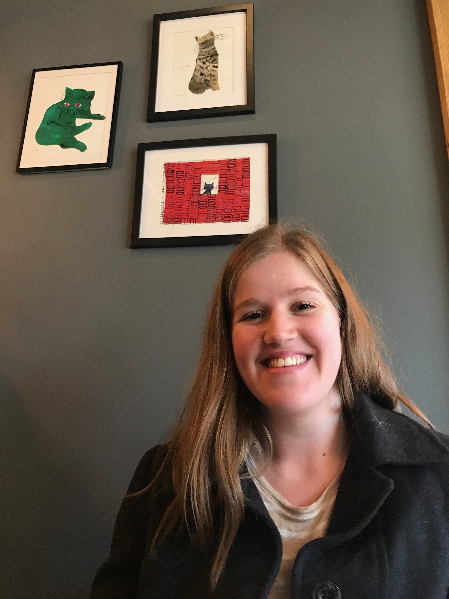 A woman with long hair smiles at the camera, seated in front of a gray wall with three framed cat artworks.