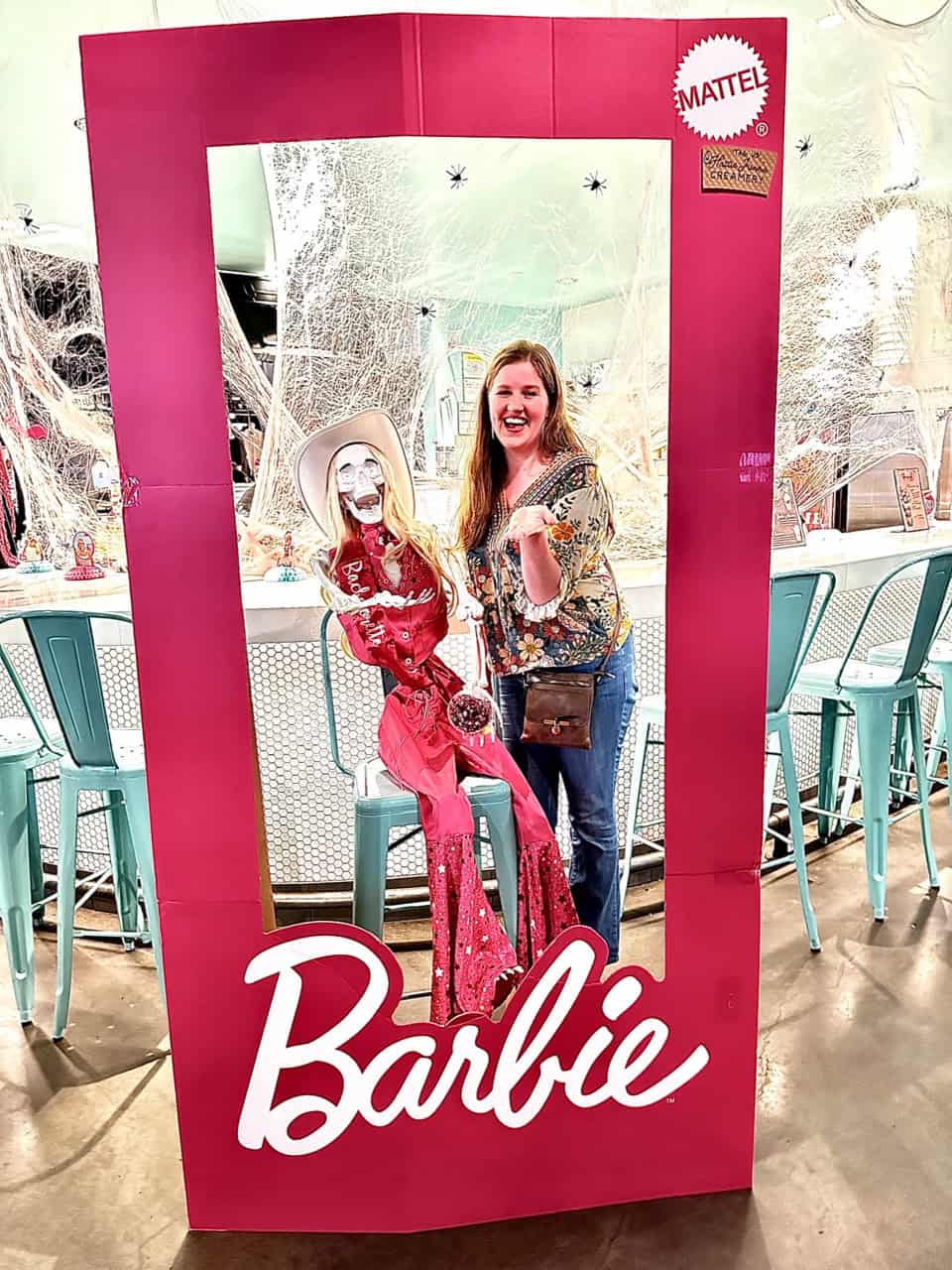A woman smiles next to a mannequin in a pink Barbie-themed display frame, set inside a stylish Nashville cafe with turquoise chairs and decorative lights.