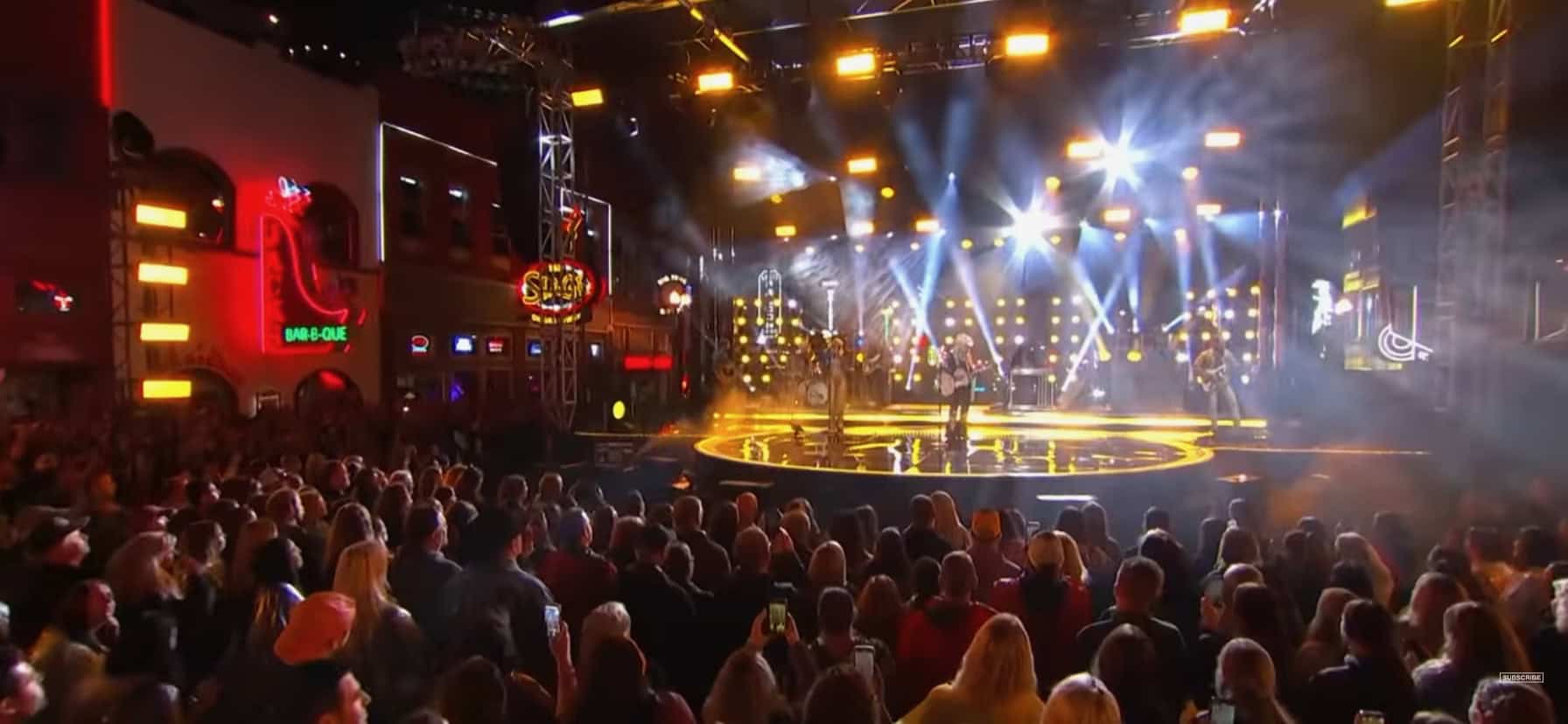 A crowd watches a lively outdoor concert on a brightly lit stage at night, surrounded by neon signs.