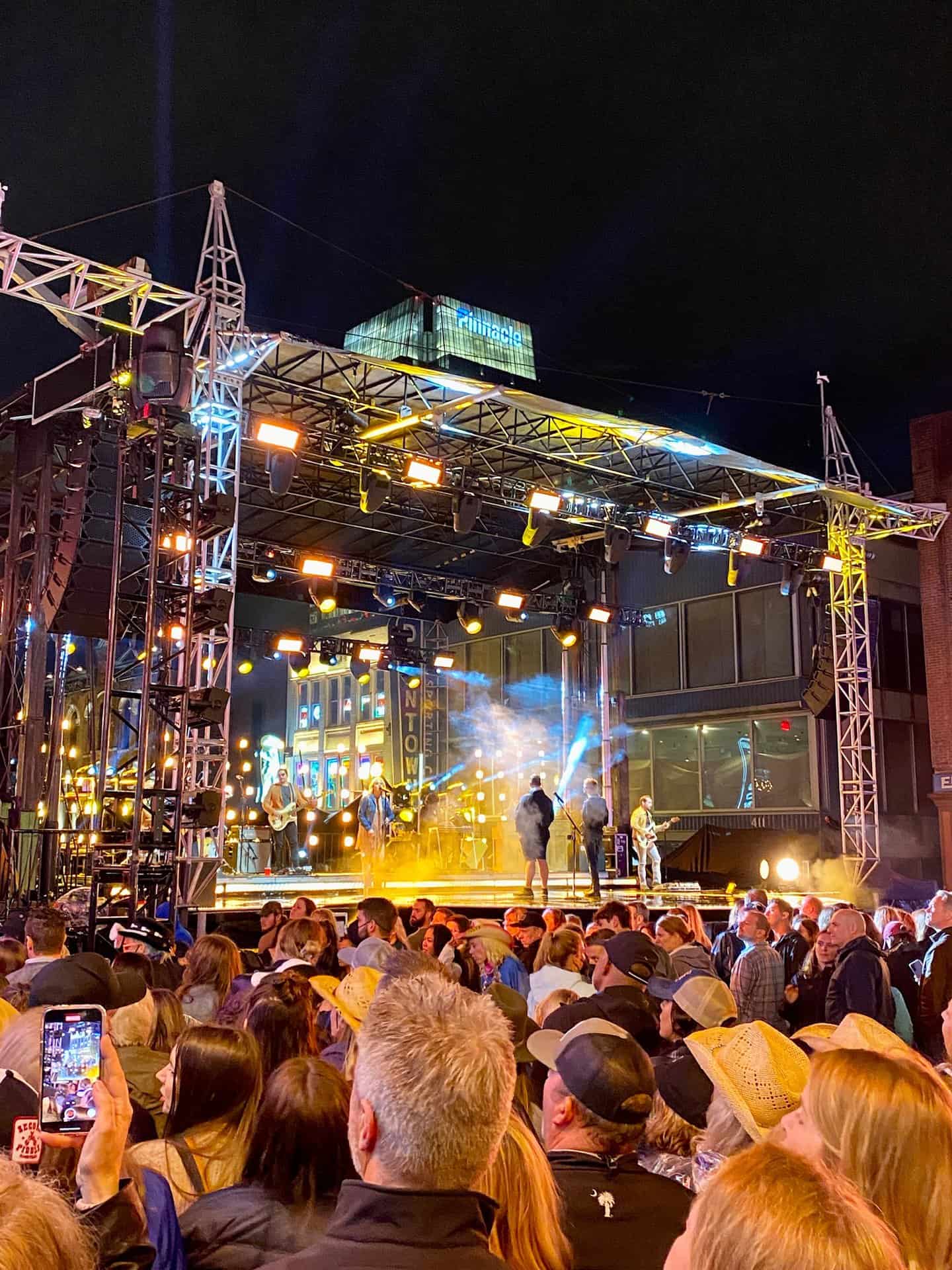 A band performs on an outdoor stage at night, surrounded by a large crowd. Lights illuminate the scene, and a building with reflective windows is in the background.