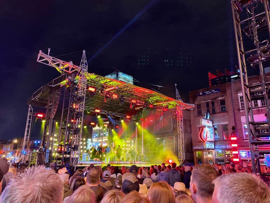 Outdoor concert at night with a crowd in front of a lit stage, featuring performers under colorful lights. Surrounding buildings are visible under the night sky.