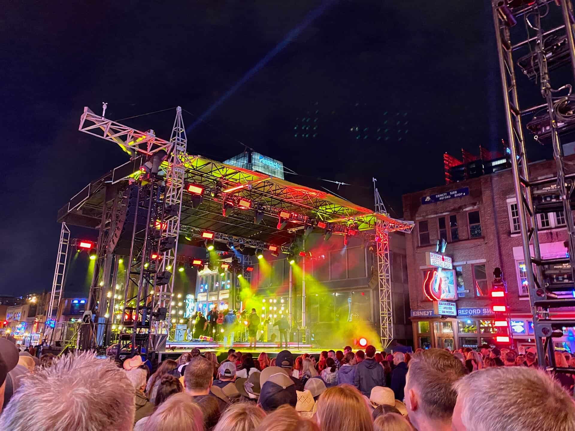 Outdoor concert at night with a crowd in front of a lit stage, featuring performers under colorful lights. Surrounding buildings are visible under the night sky.