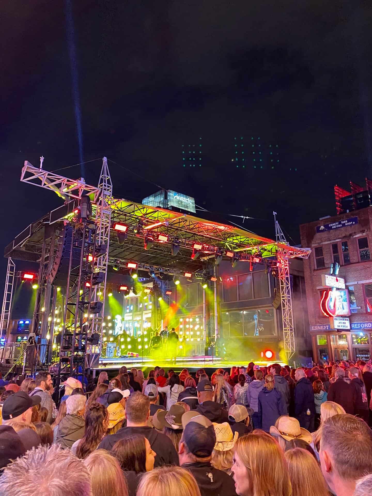Concert scene at night with a crowd watching a lit stage featuring musicians. Neon signs and buildings are visible in the background under a dark sky.