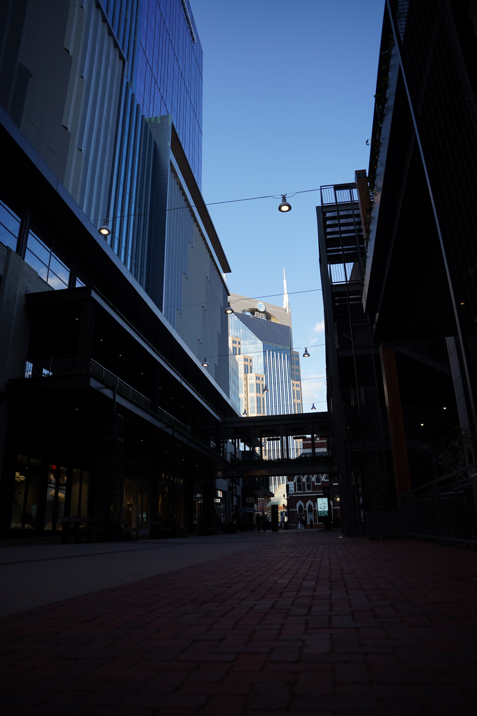 Tall modern buildings and glass facades rise above a pedestrian walkway lined with brick pavers under a clear blue sky in this vibrant Nashville city scene.
