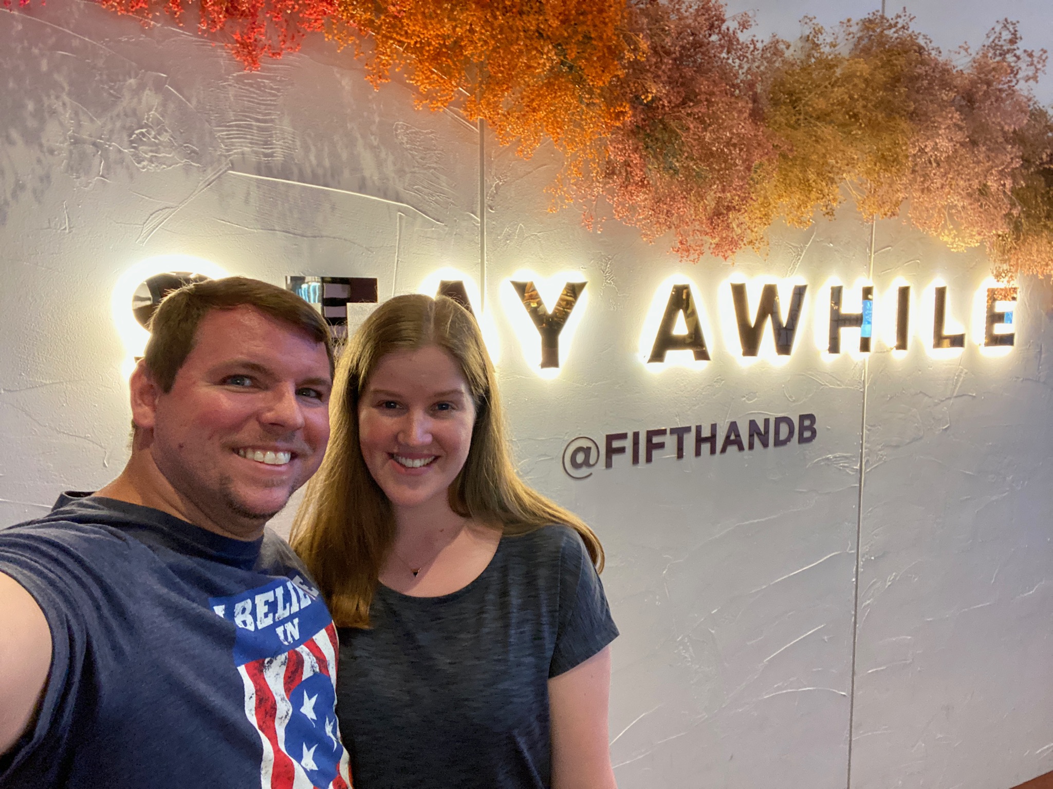 A man and woman smile in front of a wall sign that reads "STAY AWHILE @FIFTHANDB," with orange foliage decorating the top edge.