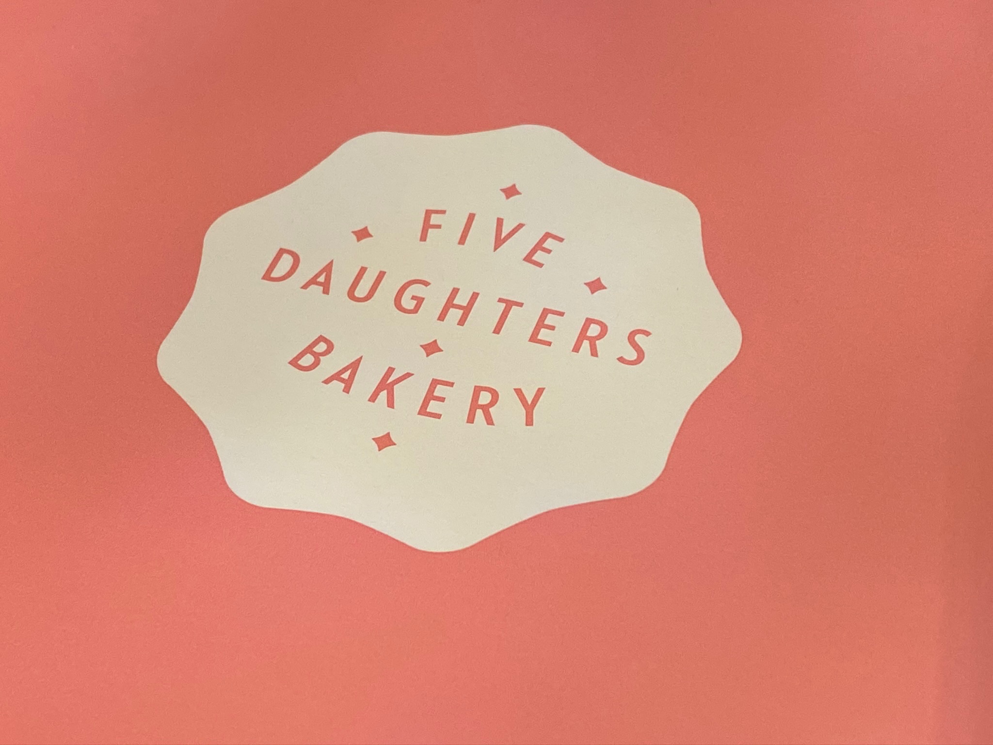 A pink background with a white, scalloped shape in the center displaying the text "FIVE DAUGHTERS BAKERY" in red capital letters.