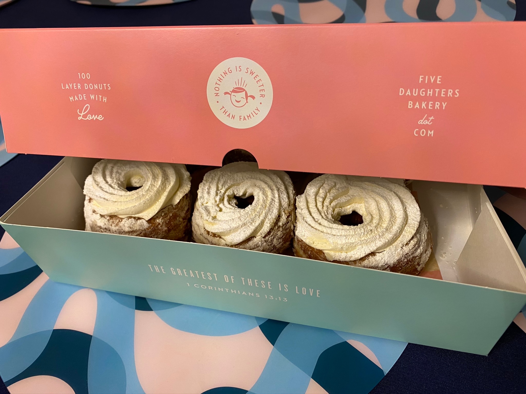 Three iced donuts in a pastel-colored box from Five Daughters Bakery, with “Nothing is sweeter than family” and a Bible verse printed on the lid and front.