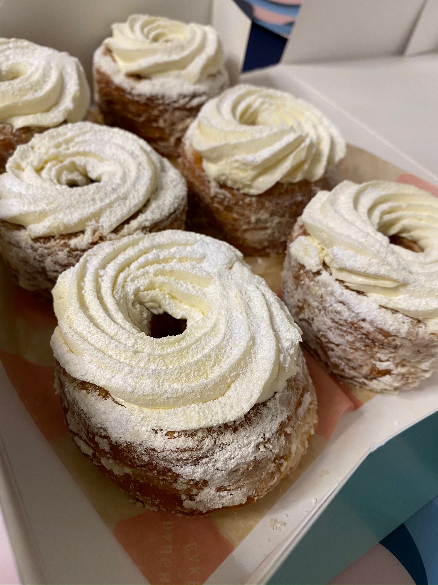 Six cronut pastries topped with swirls of cream and dusted with powdered sugar, arranged closely together in a box.
