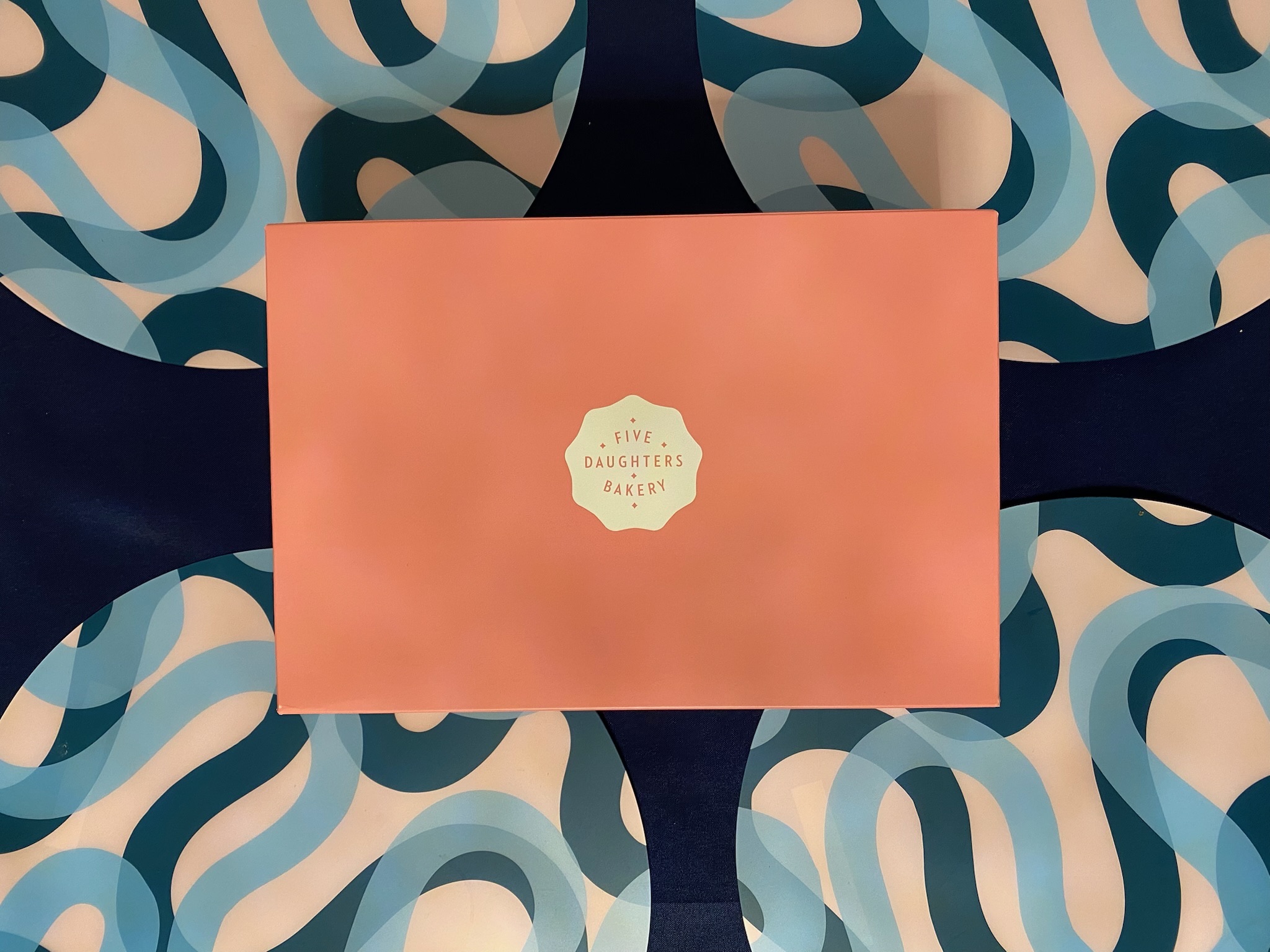 A pink Five Daughters Bakery box sits on a blue and white wavy-patterned surface.