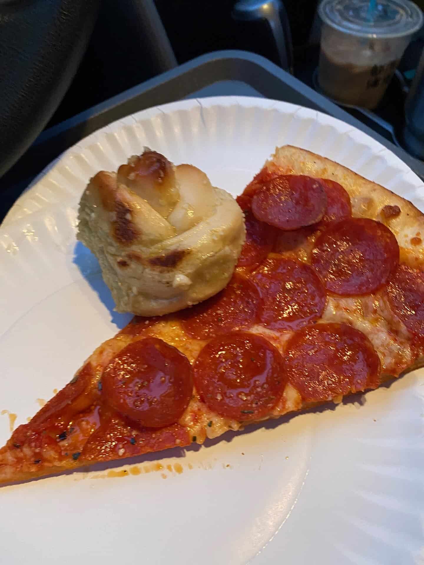 A slice of pepperoni pizza and a garlic knot on a white plate, with a plastic cup of iced coffee in the background.