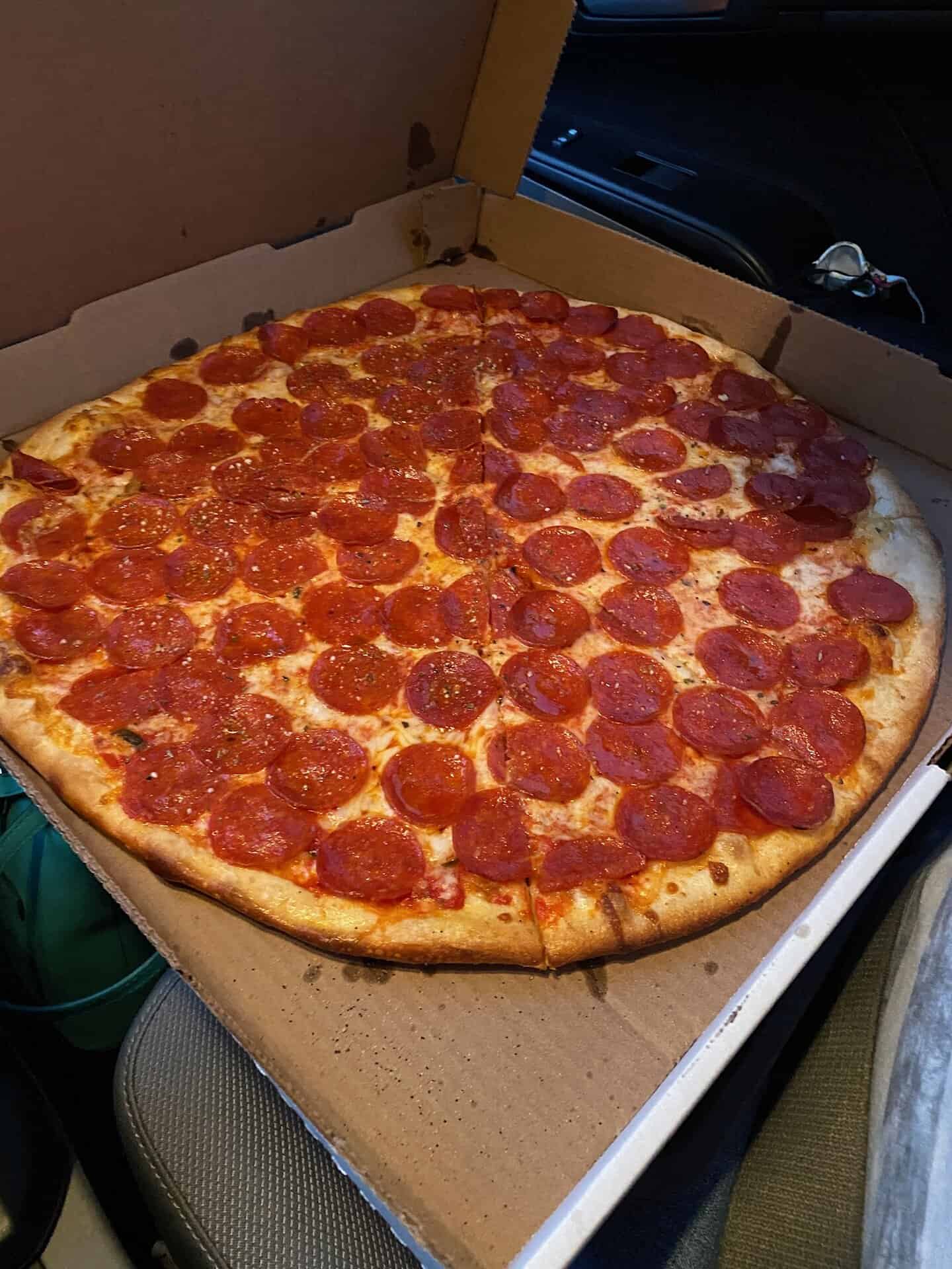 A large pepperoni pizza in a cardboard box rests on a car seat, featuring a thick crust and plentiful slices of pepperoni.