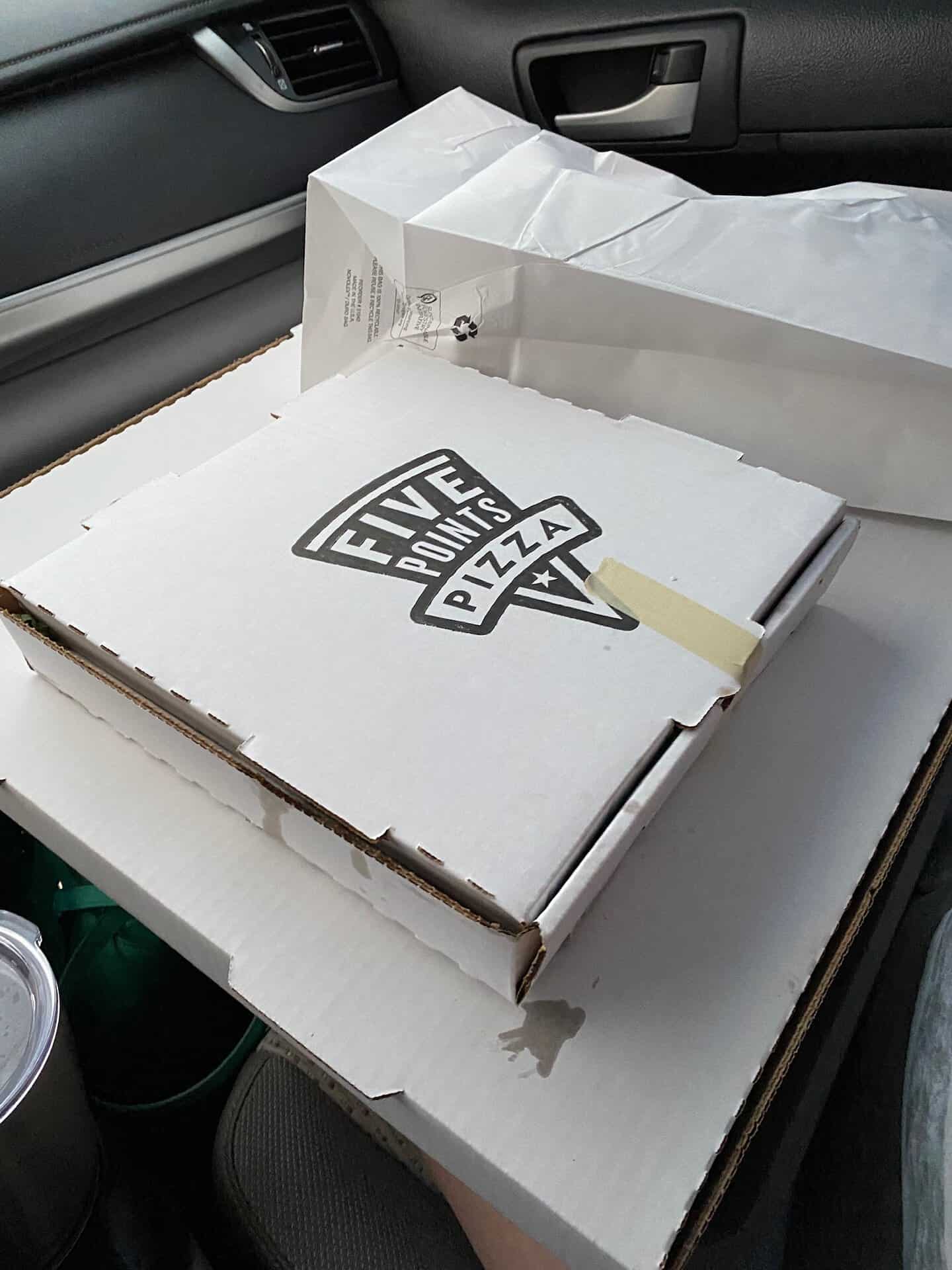 Pizza boxes and a paper bag are stacked on a car seat with a drink in the cup holder.