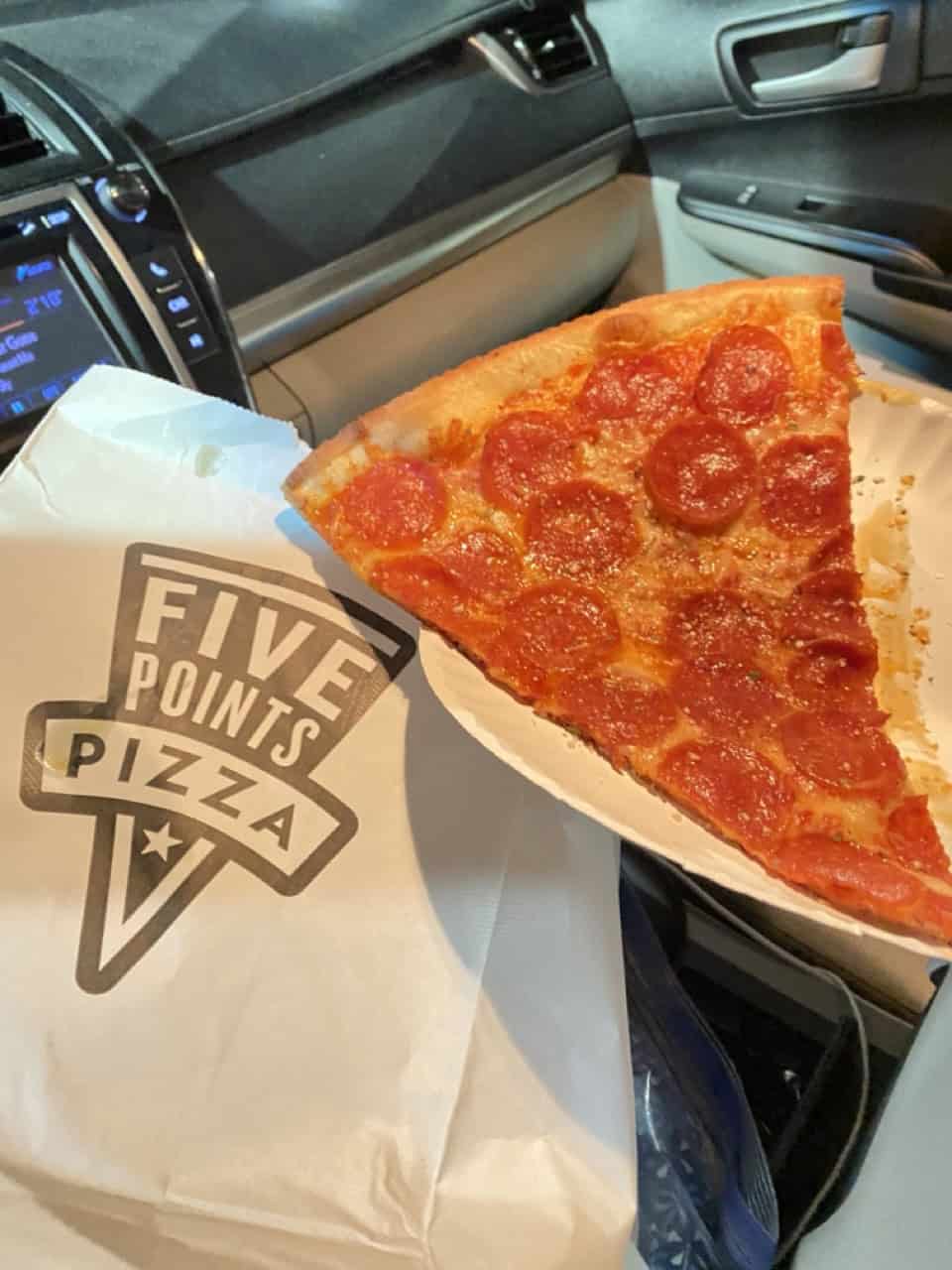 A slice of pepperoni pizza sits on a paper plate inside a car, near a bag labeled "Five Points Pizza," capturing the essence of Nashville's vibrant food scene.