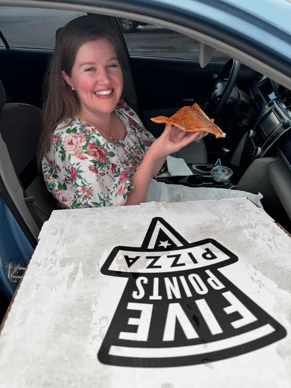 A person sits in a car, smiling with a slice of pizza in hand, embracing the flavorful joy of Nashville's own "Five Points Pizza." An open box from this iconic spot rests in the foreground, promising more deliciousness to savor.