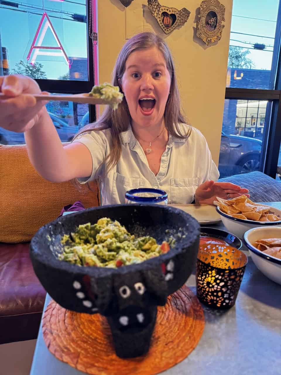 Person excitedly holding a spoonful of guacamole from a large stone bowl on a dining table, with candles and other dishes nearby, ready to savor the flavors of this Nashville-inspired feast.