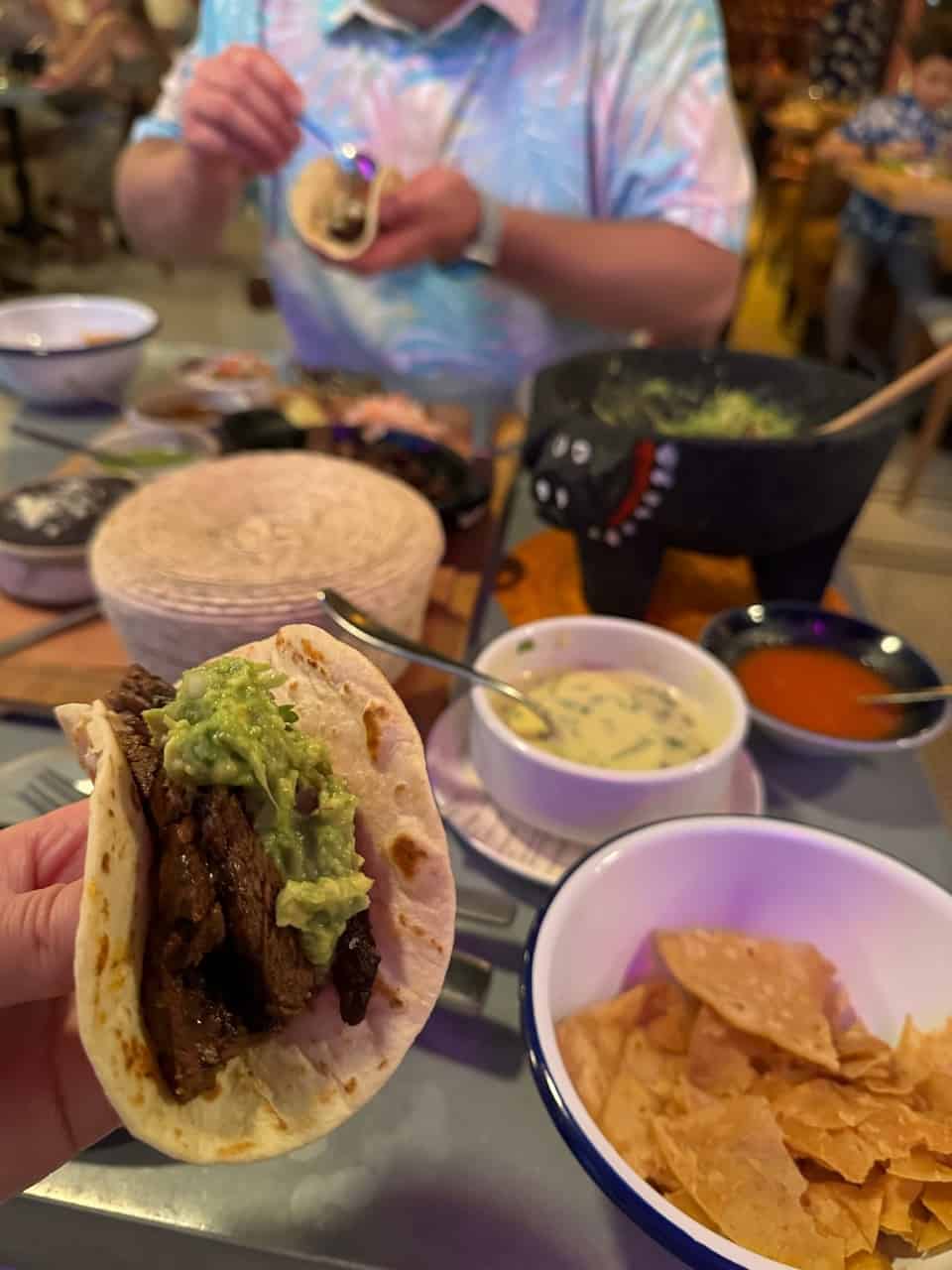 A person holds a taco overflowing with meat and guacamole, while the table is laden with bowls of chips, sauce, and tortillas. In the background, another culinary enthusiast prepares food, adding a hint of Nashville flair to the vibrant scene.