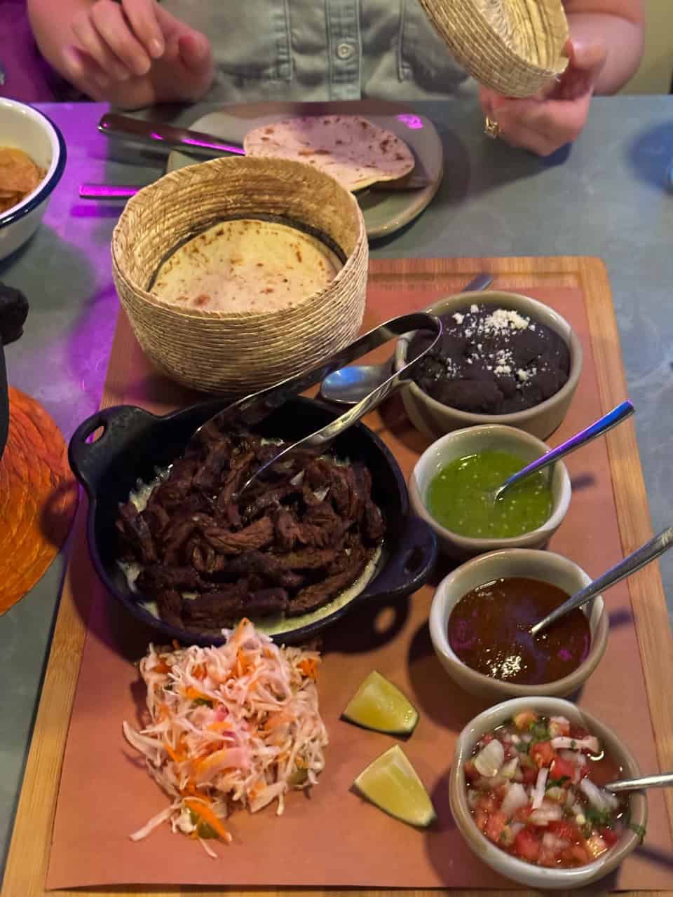 A wooden board showcases Nashville-inspired tacos, grilled beef, coleslaw, lime wedges, and bowls of guacamole, salsa, black beans, and pico de gallo. In the background, a person holds an empty tortilla basket.