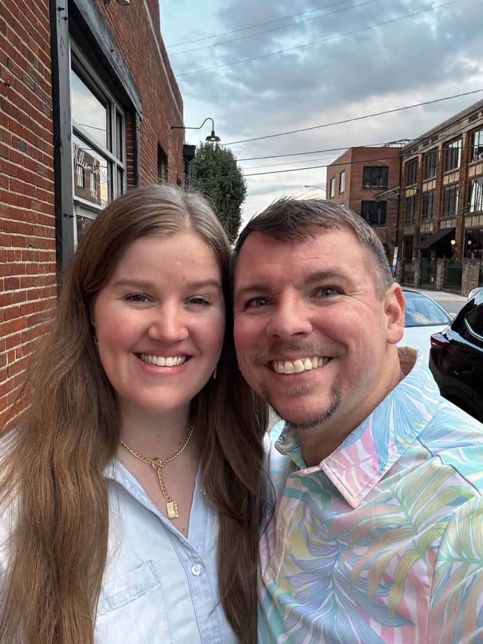 A man and woman are smiling, standing outside with a brick building backdrop in the heart of Nashville, surrounded by parked cars and a cloudy sky.