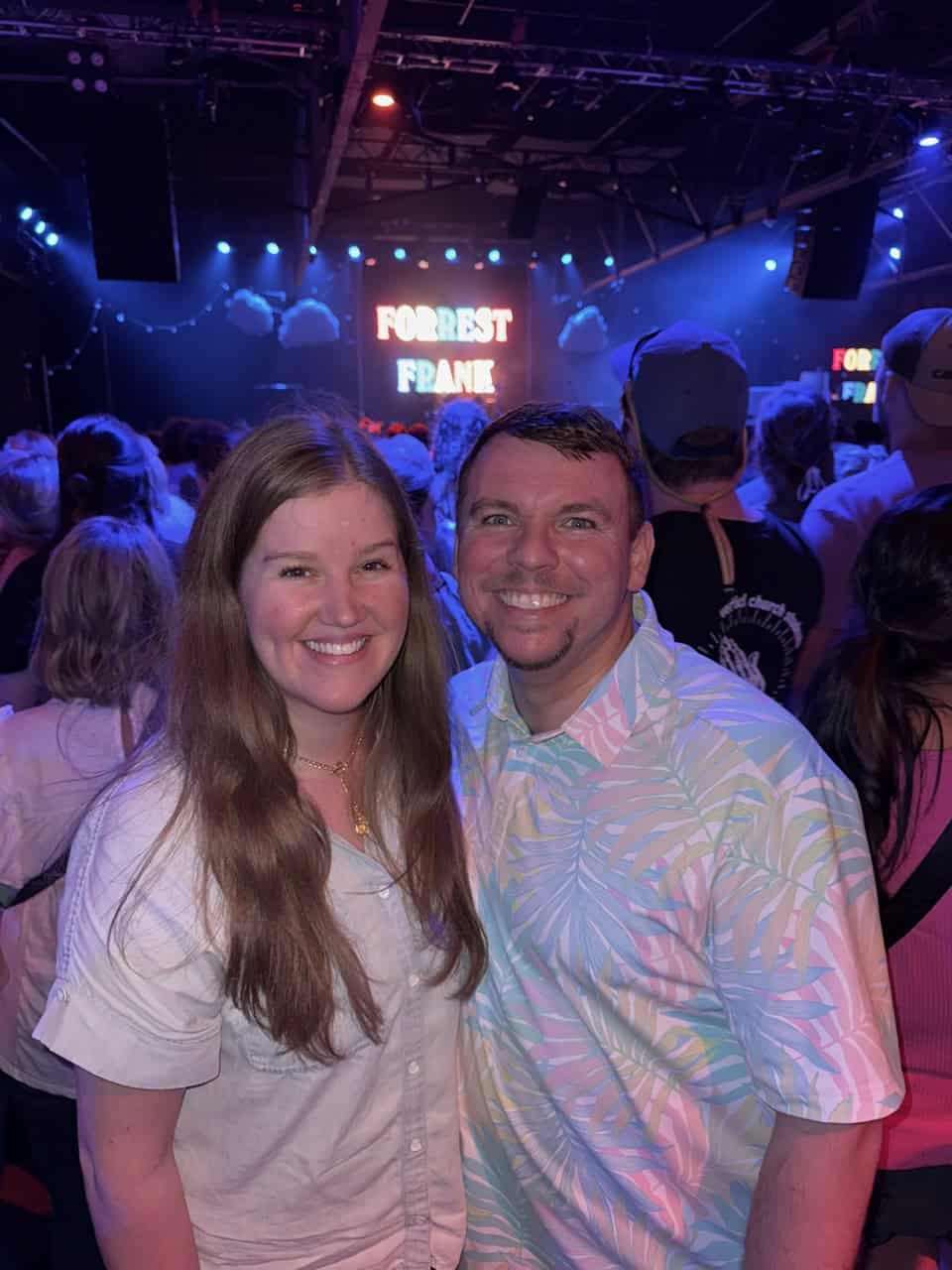 A man and woman smiling at a concert venue in Nashville, with a crowd in the background and a sign that reads "Forest Frank.