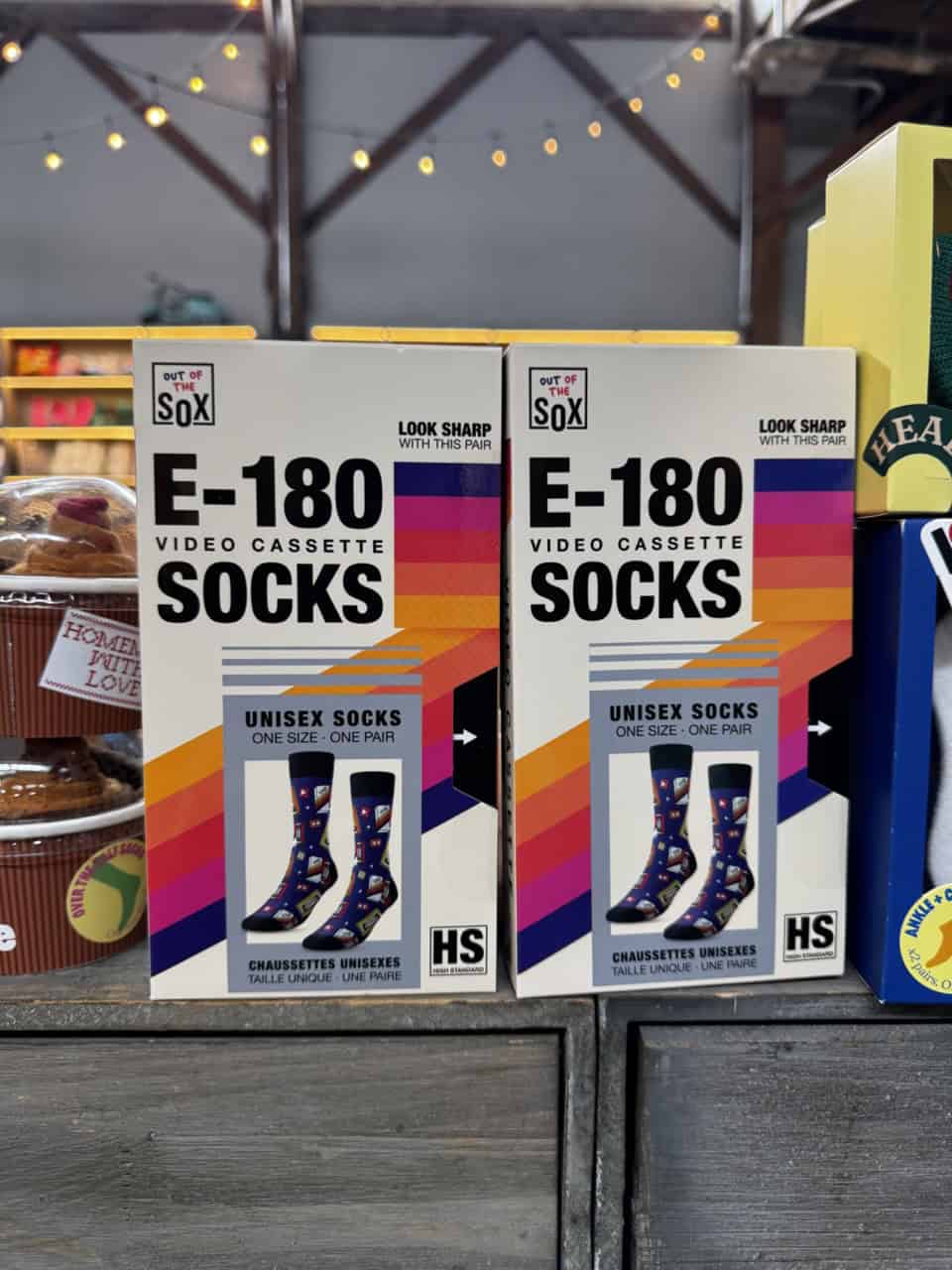 Two boxes of E-180 Socks with a retro VCR tape design sit on a Nashville store shelf. The packaging features colorful sock illustrations, describing them as unisex and one size fits most.