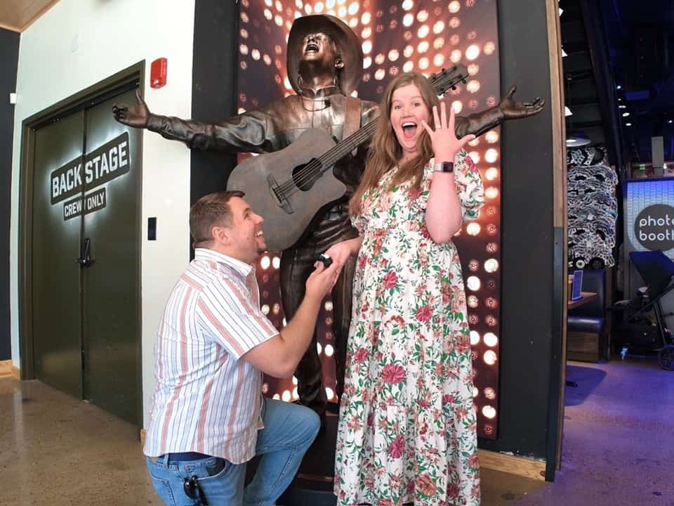 In Nashville, a man kneels before a surprised woman in front of a statue with a guitar, capturing the magic of Music City. She beams as she displays the ring on her hand.