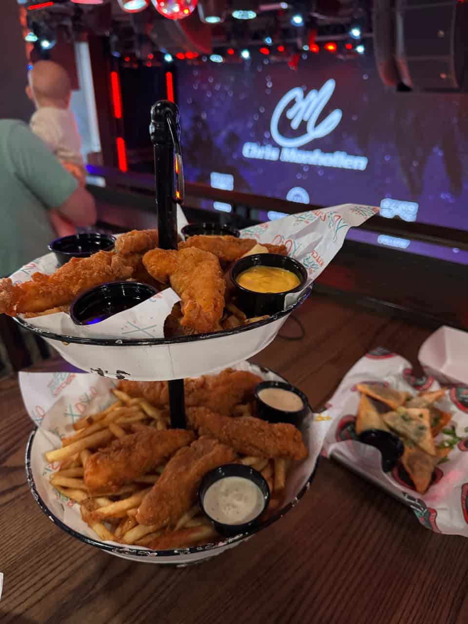 Two-tiered platter with Nashville-style fried chicken strips, fries, and dips. A plate with pizza slices on the side adds to the feast. The dimly lit venue hums with energy, a vibrant crowd mingles as a stage bears a visible logo in the background.