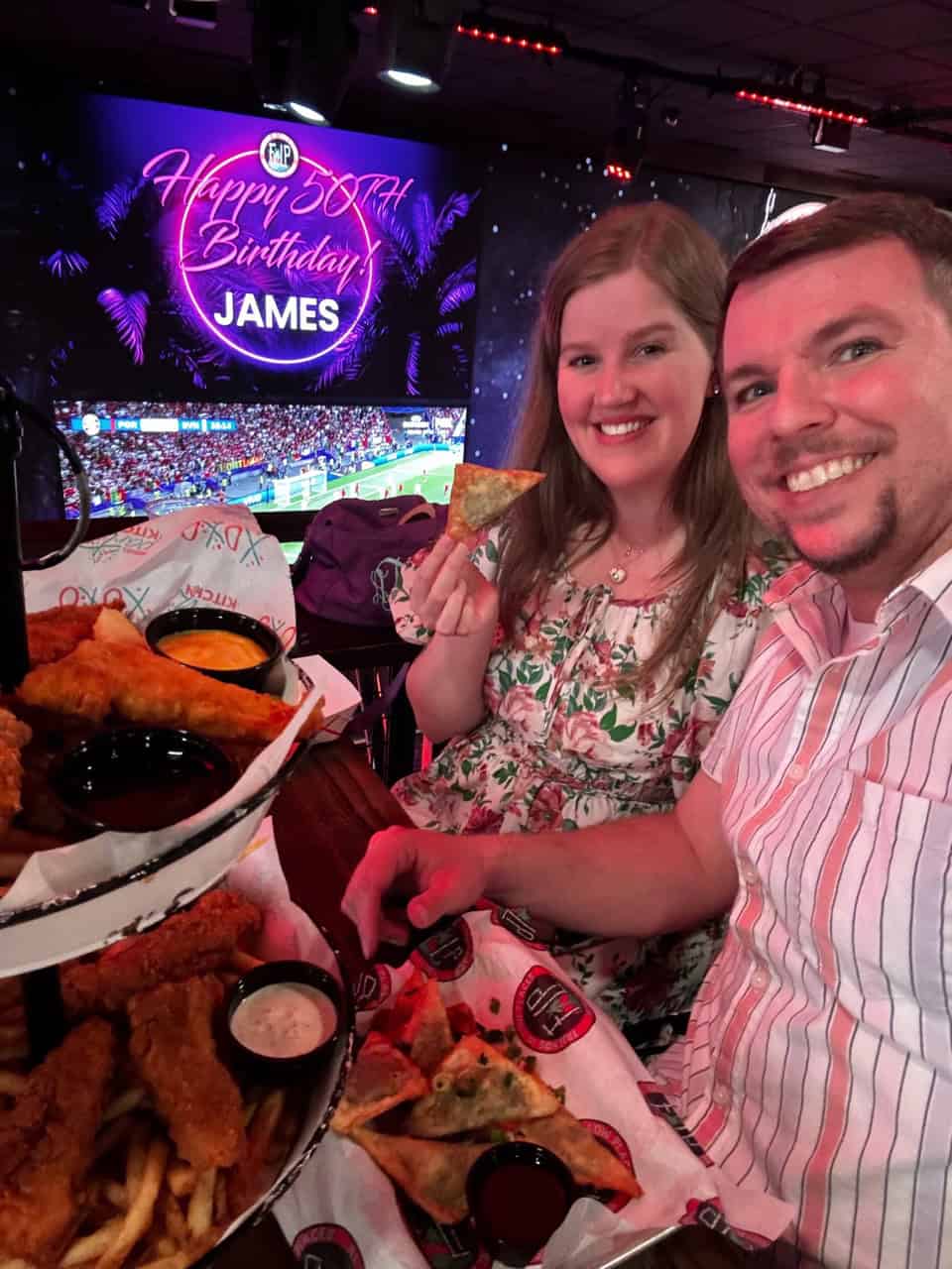 A man and woman smile at a table with fried food, capturing the spirit of Nashville's lively charm. Behind them, a screen proudly displays "Happy 30th Birthday James" while a thrilling sports event unfolds.