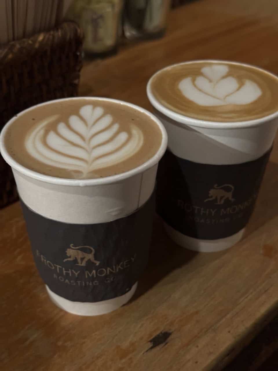 Two cups of coffee with latte art sit on a wooden table in a cozy Nashville café. Both cups have Frothy Monkey sleeves, with one featuring a leaf design and the other a simple swirl.