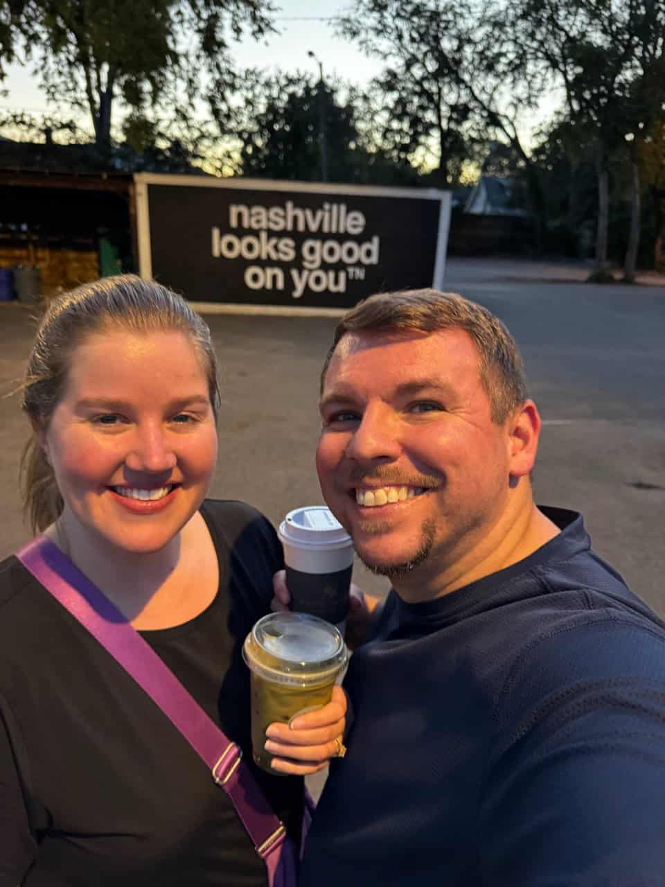 Two people smiling, holding drinks outside with a sign in the background proudly declaring, "Nashville looks good on you." Trees and a building are bathed in the warm evening light, perfectly capturing the charm of Nashville.
