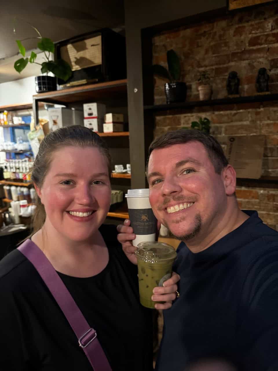 A man and woman smile while holding drinks in a charming Nashville coffee shop with a brick wall and shelves in the background.