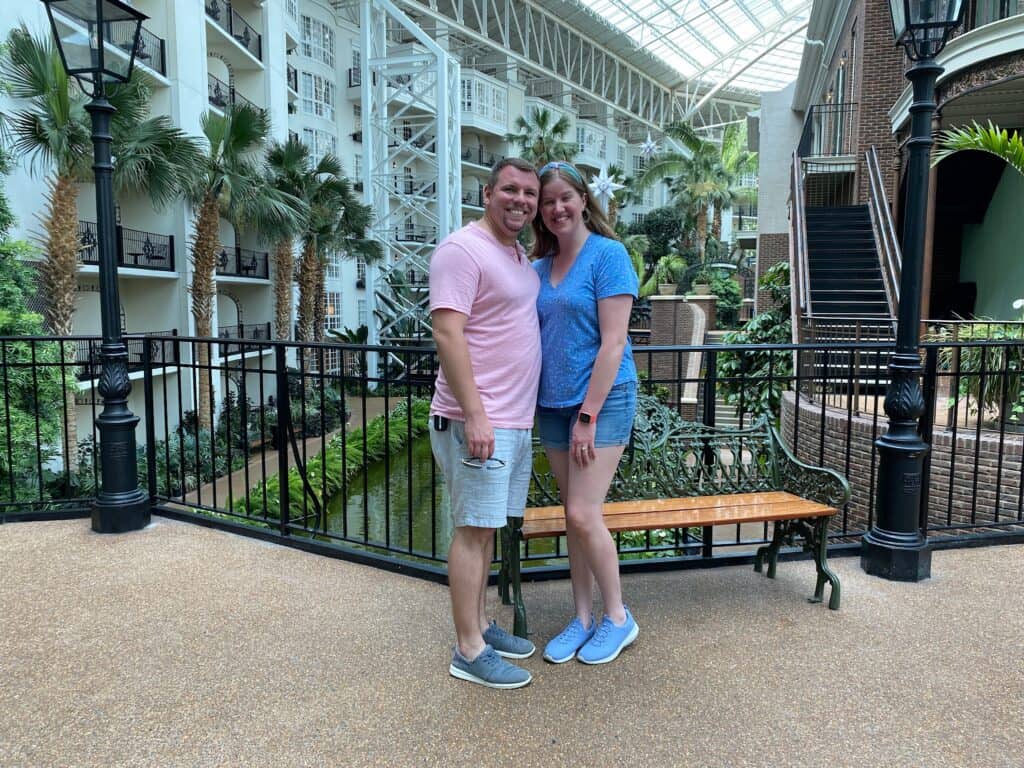 Two people stand smiling in an indoor setting with palm trees, white buildings, and a glass ceiling. They are standing near a wooden bench.