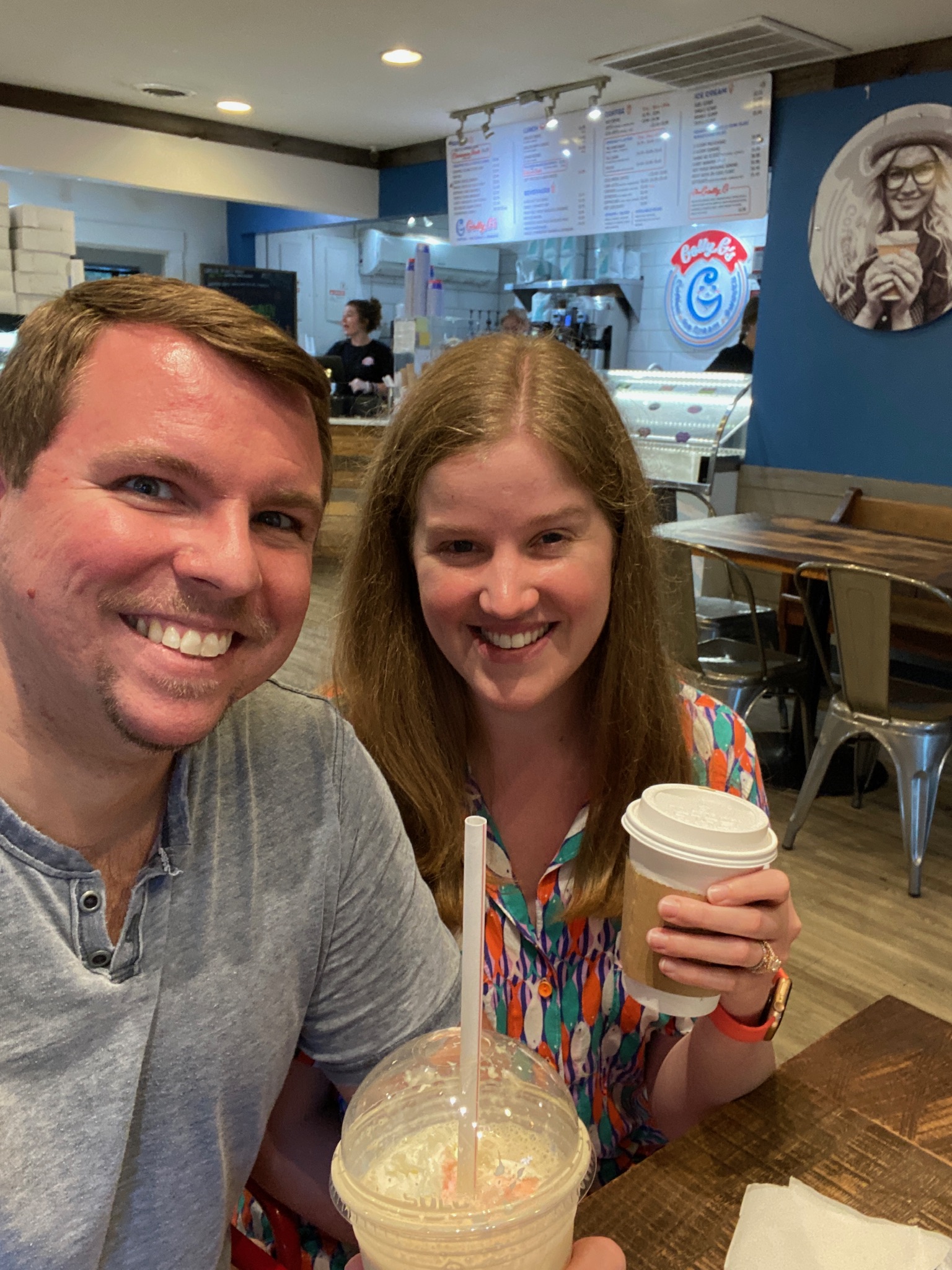 A man and woman sit at a table in a cafe, smiling at the camera while holding drinks; the cafe interior is visible in the background.