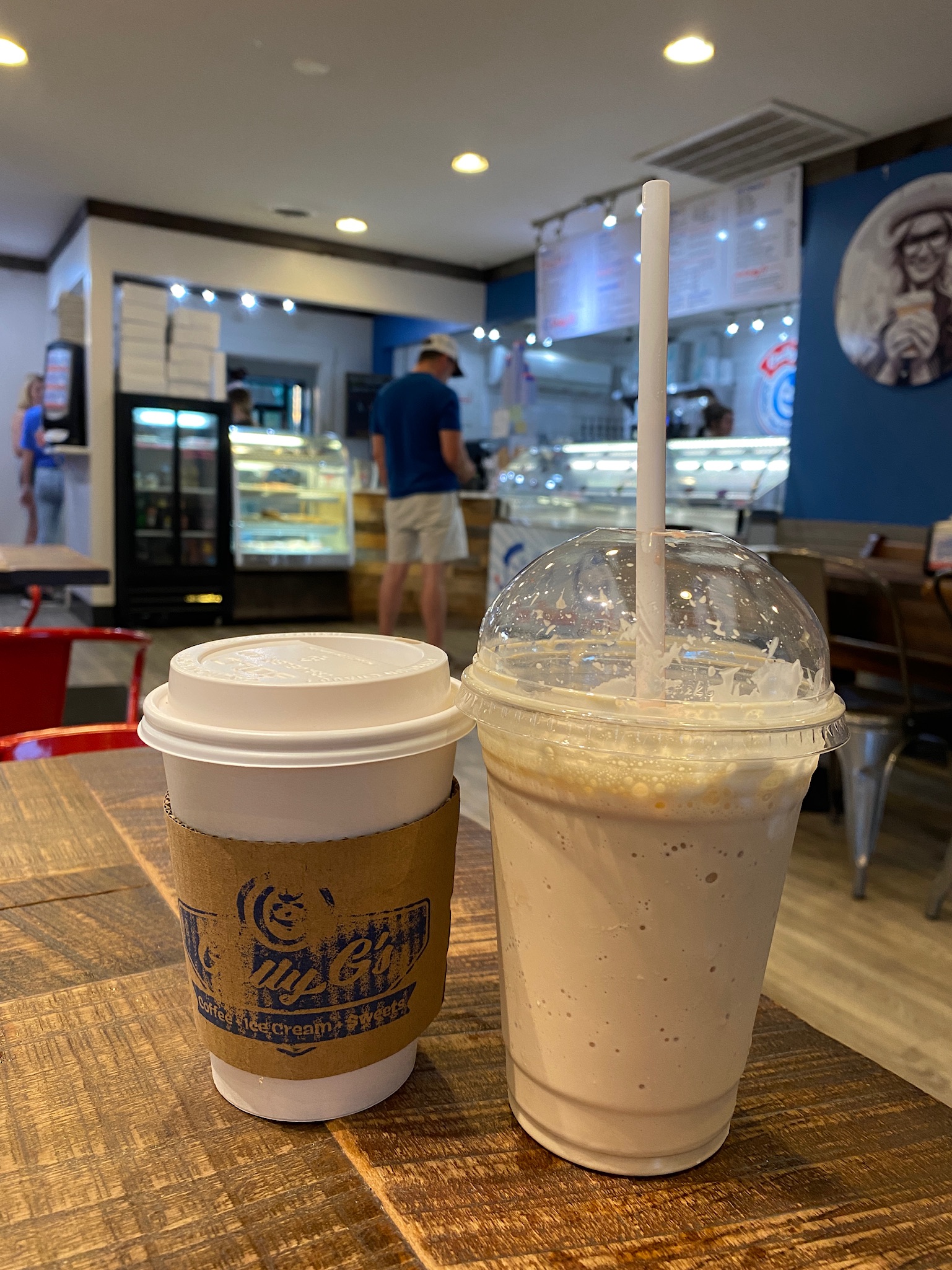 A hot coffee in a takeaway cup and an iced blended drink with a straw sit on a wooden table inside a casual café with a counter and menu in the background.