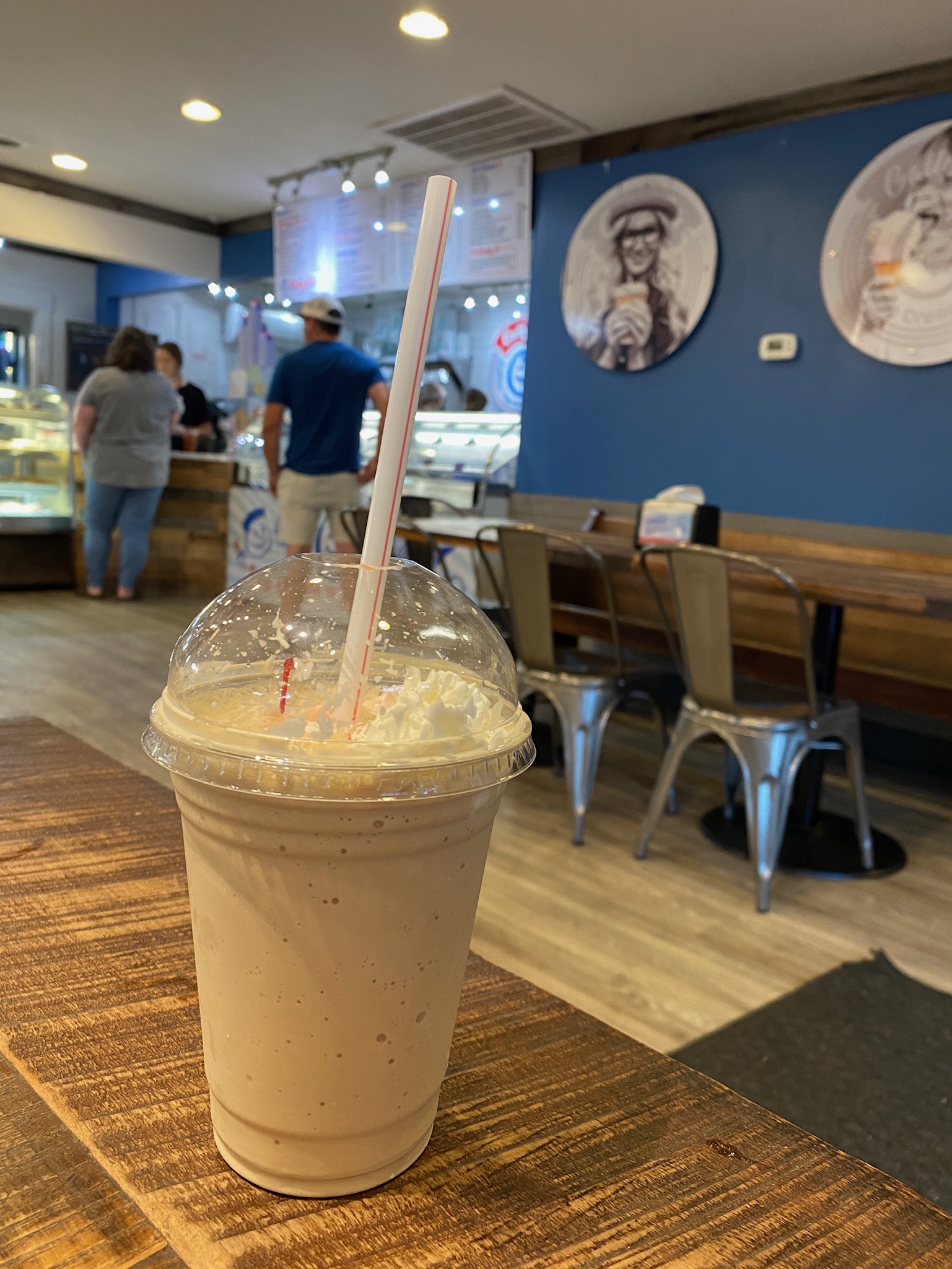 A vanilla milkshake with whipped cream sits on a wooden table in a casual ice cream shop with people ordering at the counter in the background.