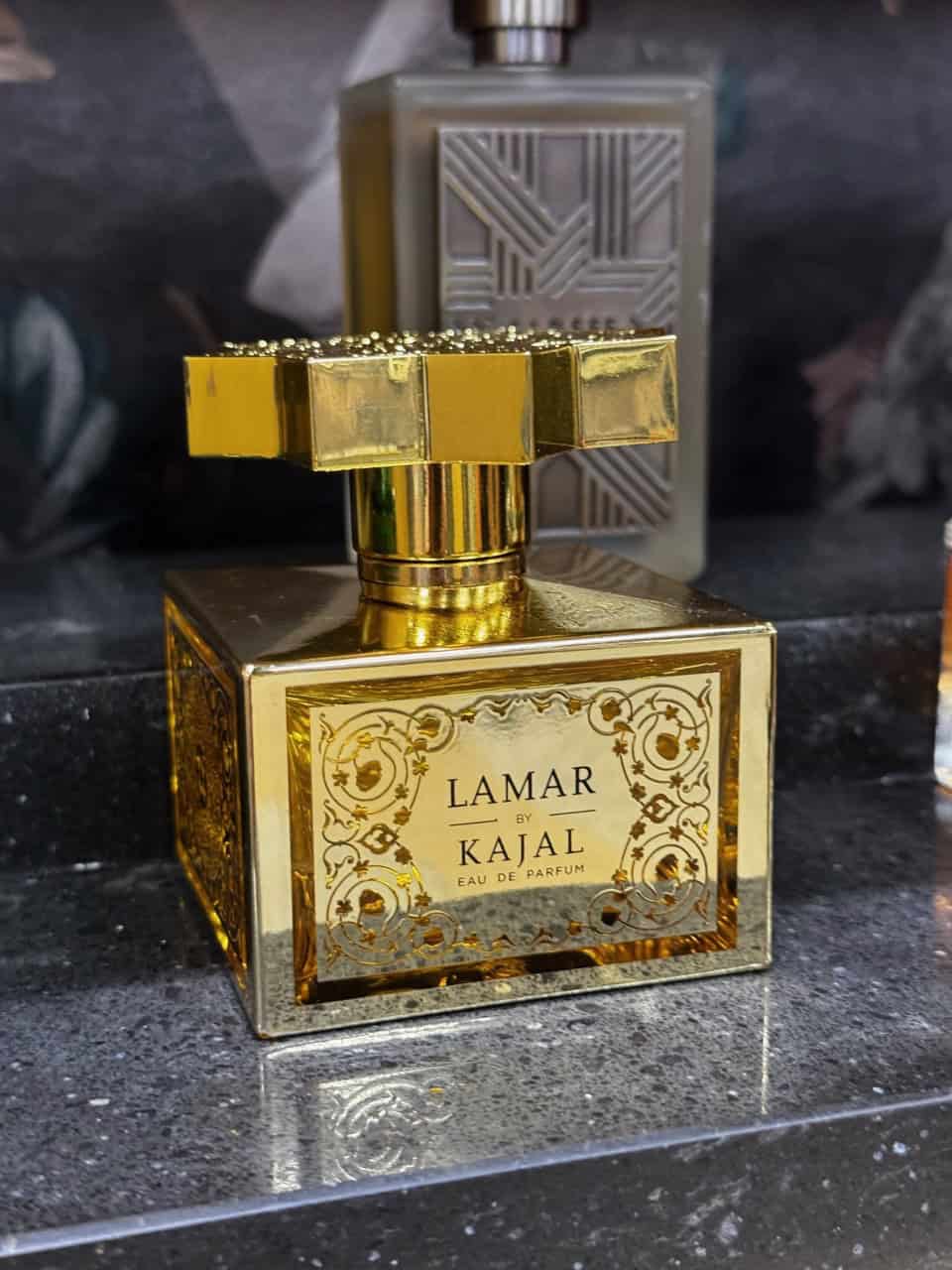 A gold-colored perfume bottle labeled "Lamar by Kajal, Eau de Parfum" rests on a speckled gray surface, reminiscent of Nashville’s timeless charm, with a larger rectangular bottle in the background.