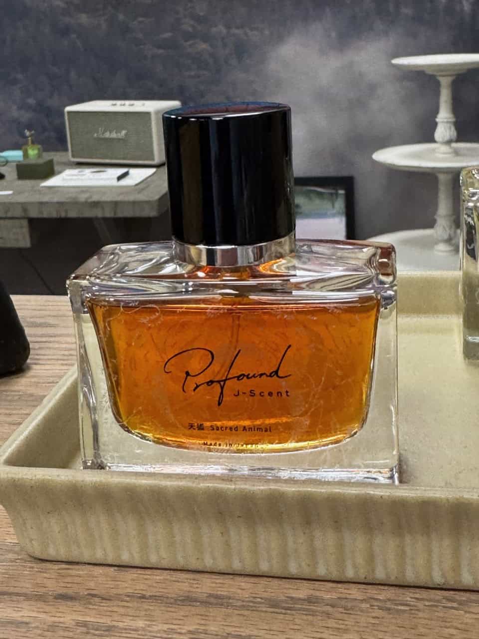 A bottle of "Profound J-Scent" perfume with amber liquid rests on a beige tray, evoking the rustic charm of Nashville. In the background, shelves adorned with a speaker and decor items complete the scene.