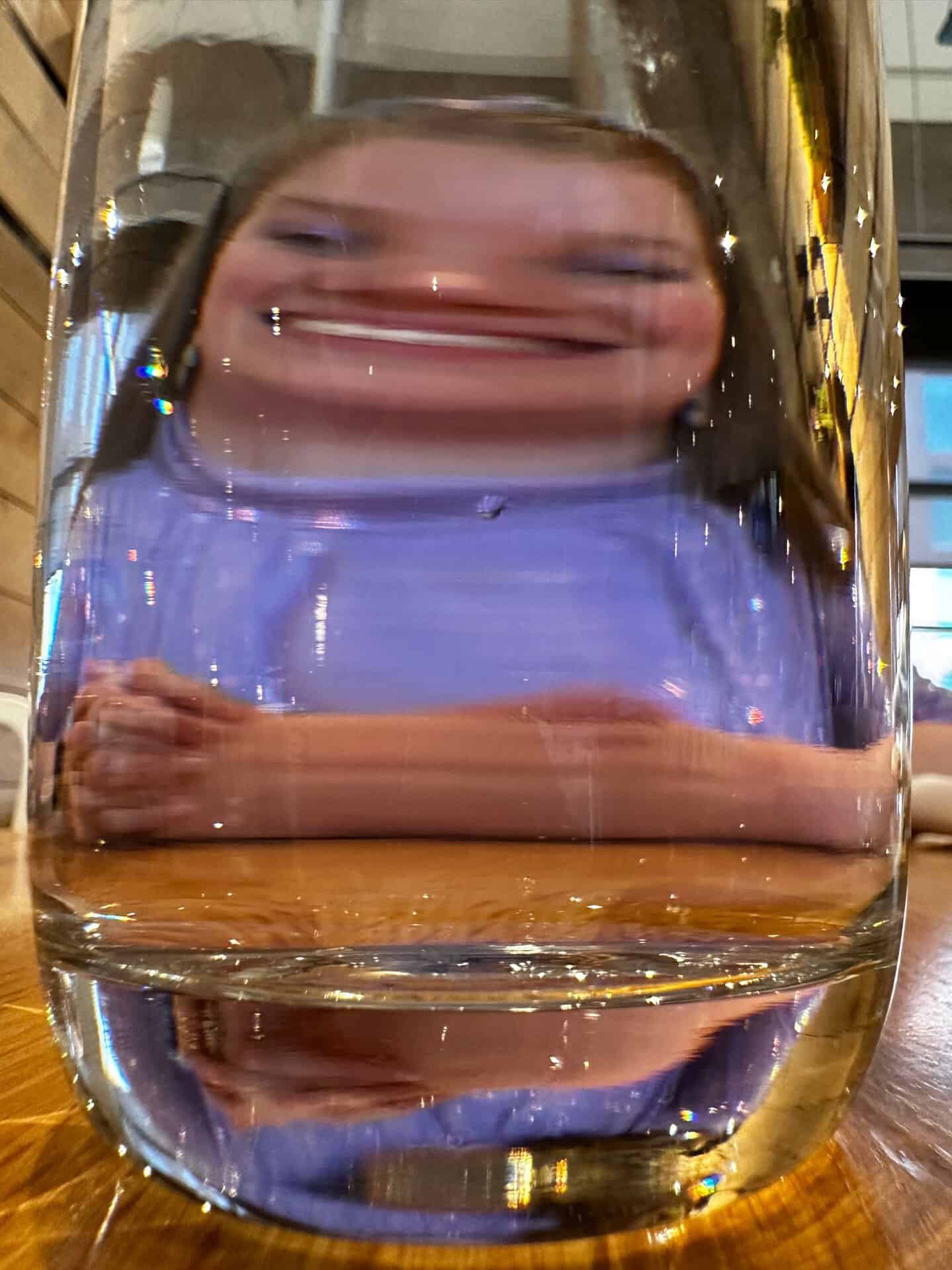 A woman's face, humorously distorted by a glass of water on a wooden table in Nashville, features her hands visible in the playful scene.