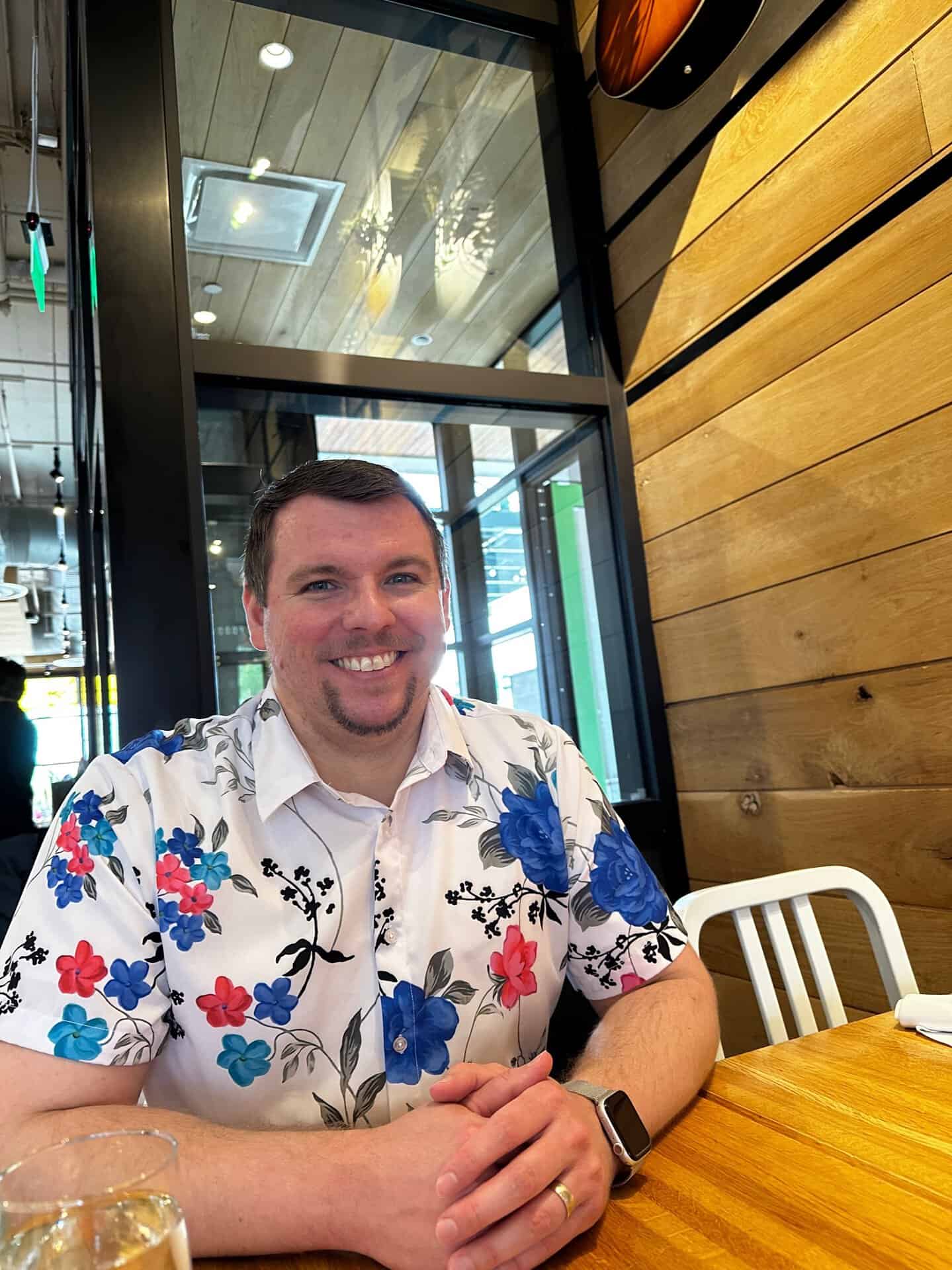 A man in a floral shirt sits at a wooden table, glowing with the charm of Nashville, in a modern restaurant adorned with sleek glass and warm wood decor, smiling brightly at the camera.