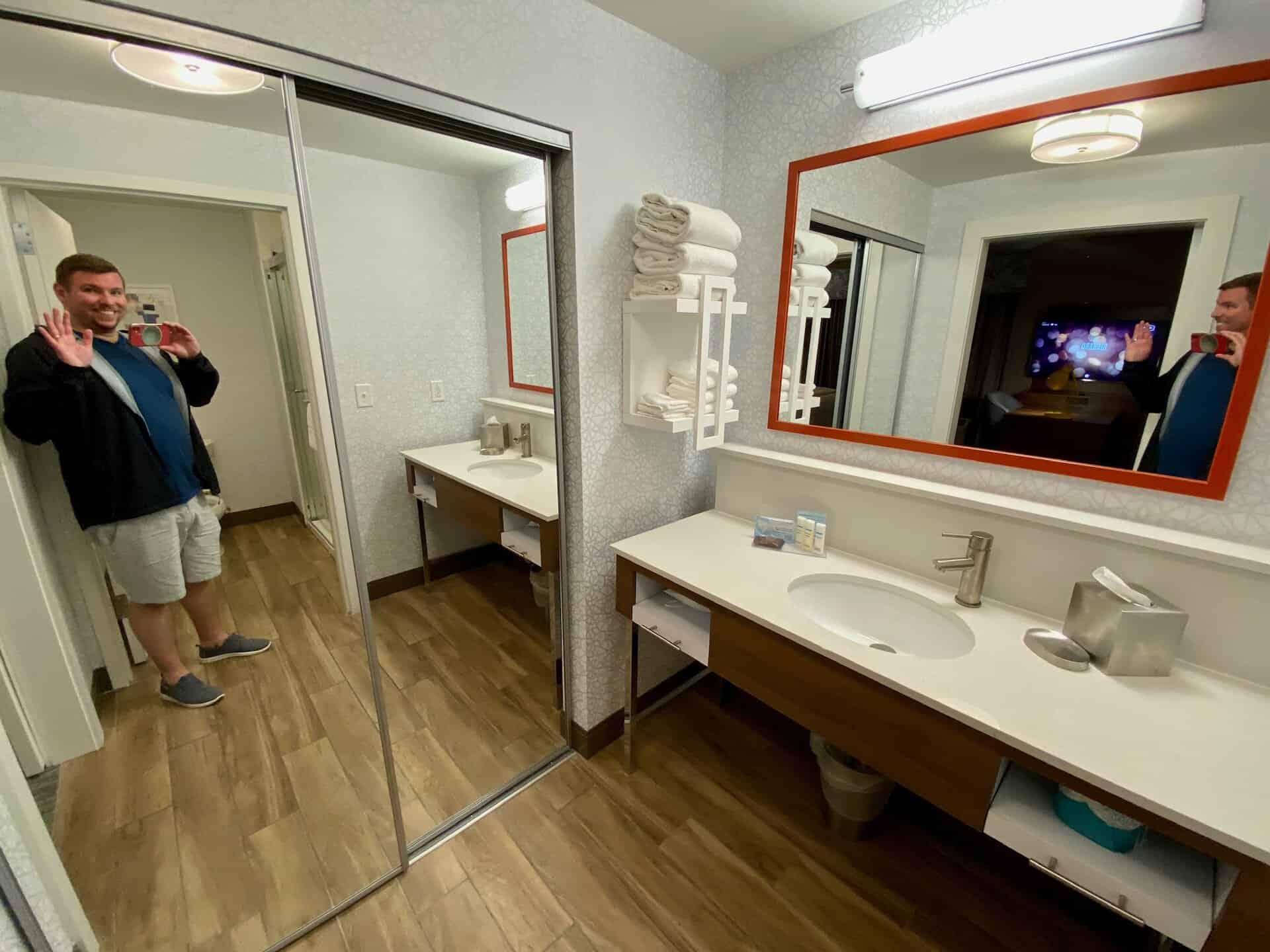A man waves in a bathroom with a mirrored closet, single sink vanity, clean towels, and a visible TV screen reflection.