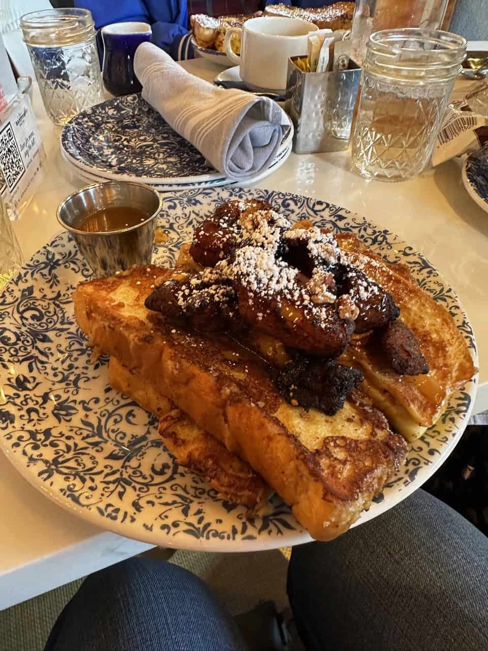 Plate of French toast topped with powdered sugar and sliced bananas, served Nashville-style with syrup in a small cup, on a patterned plate.