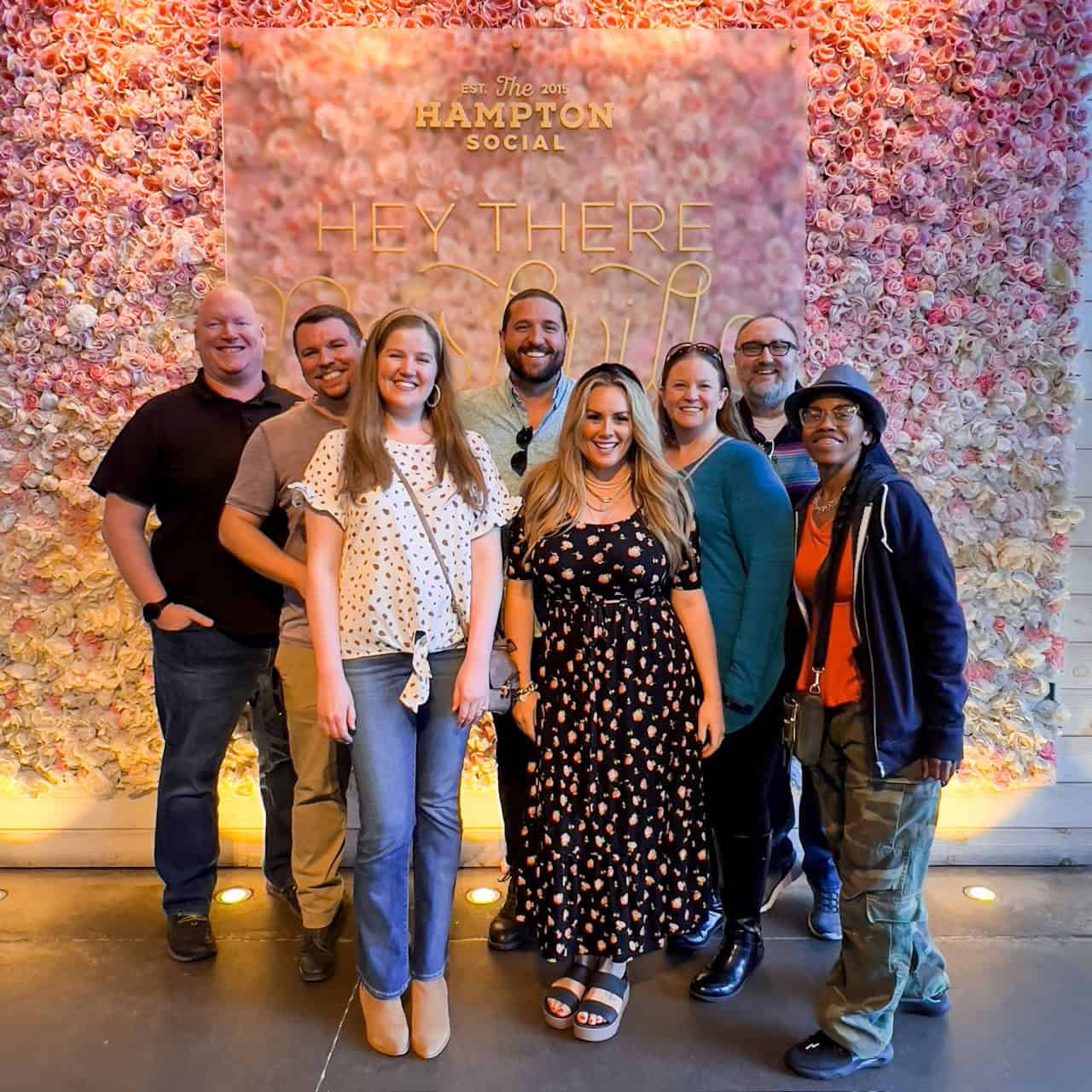 A group of eight people enjoys the moment in front of a stunning floral wall display at The Hampton Social, Nashville, cheerfully posing beneath the text "Hey There.