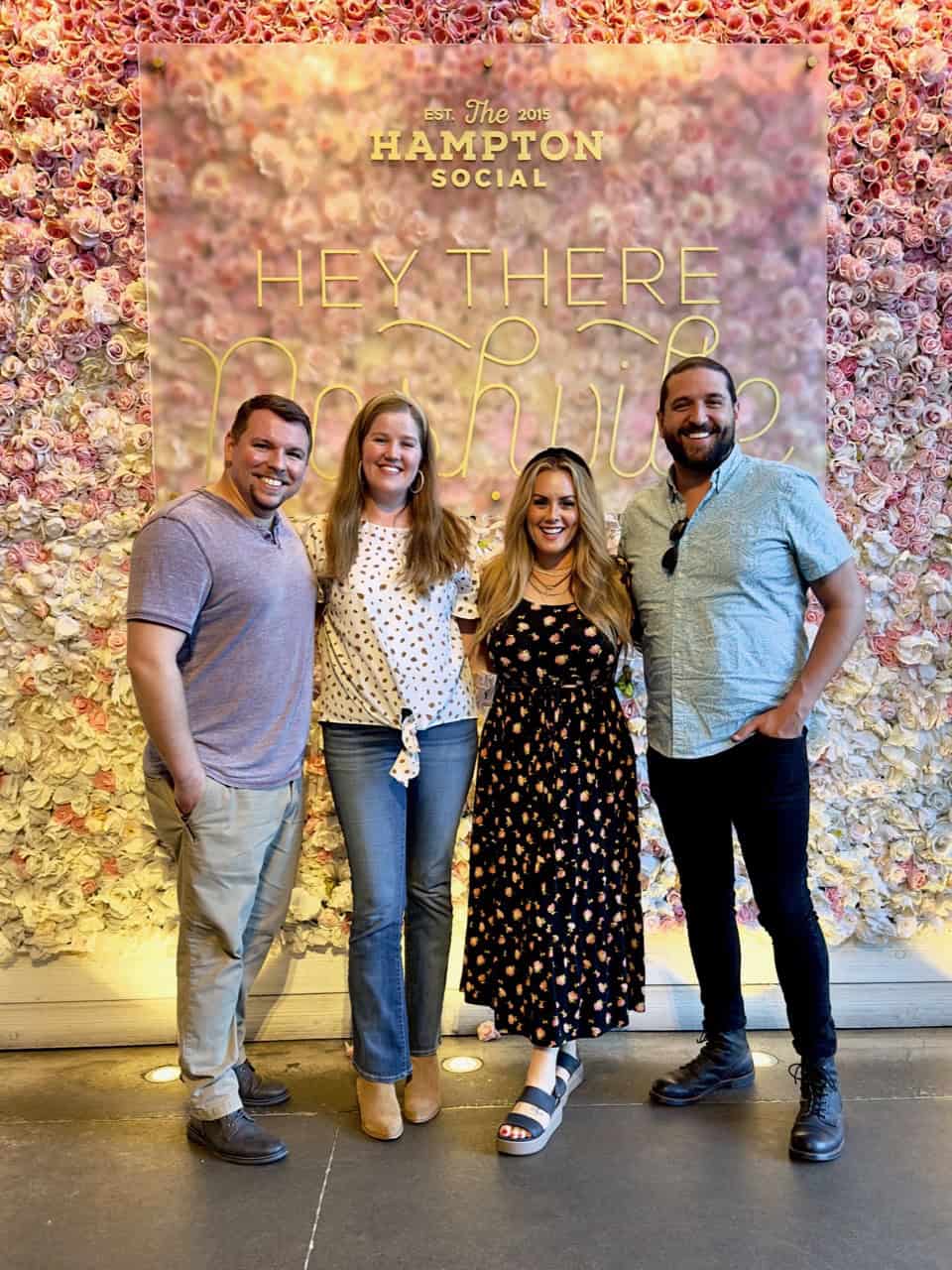 Four people stand smiling in front of a vibrant floral wall display at The Hampton Social, capturing the lively spirit of Nashville.