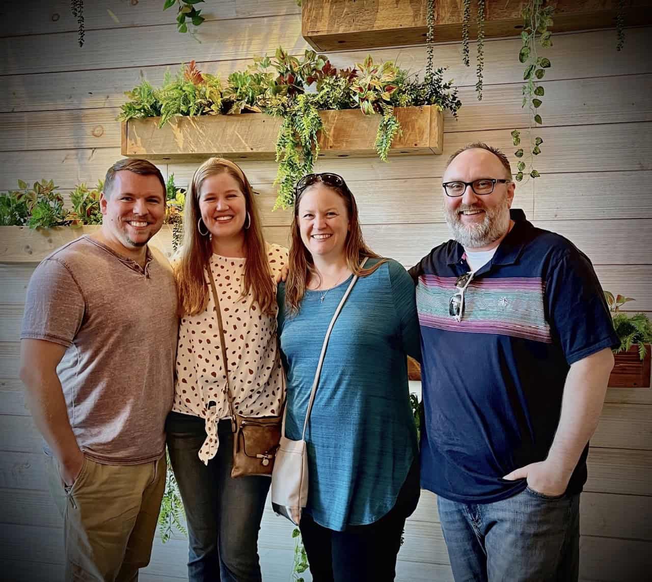 Four people standing in front of a wooden wall adorned with hanging plants, smiling at the camera, capture the vibrant essence of Nashville.